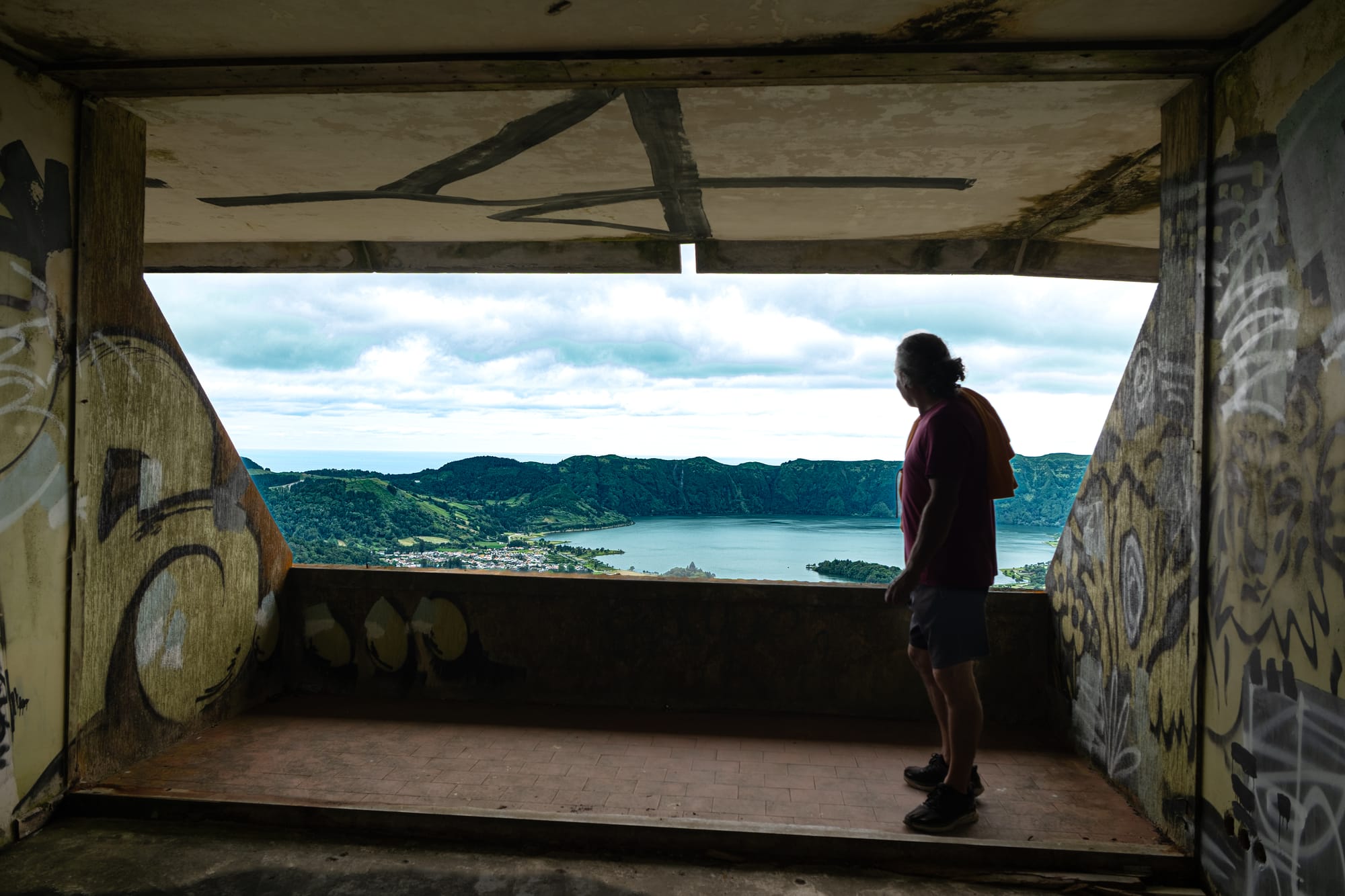 Interior of an abandoned hotel room with graffiti on the walls and ceiling, showing a man standing near a wide opening that overlooks the lakes of Sete Cidades and surrounding green ridges under a cloudy sky