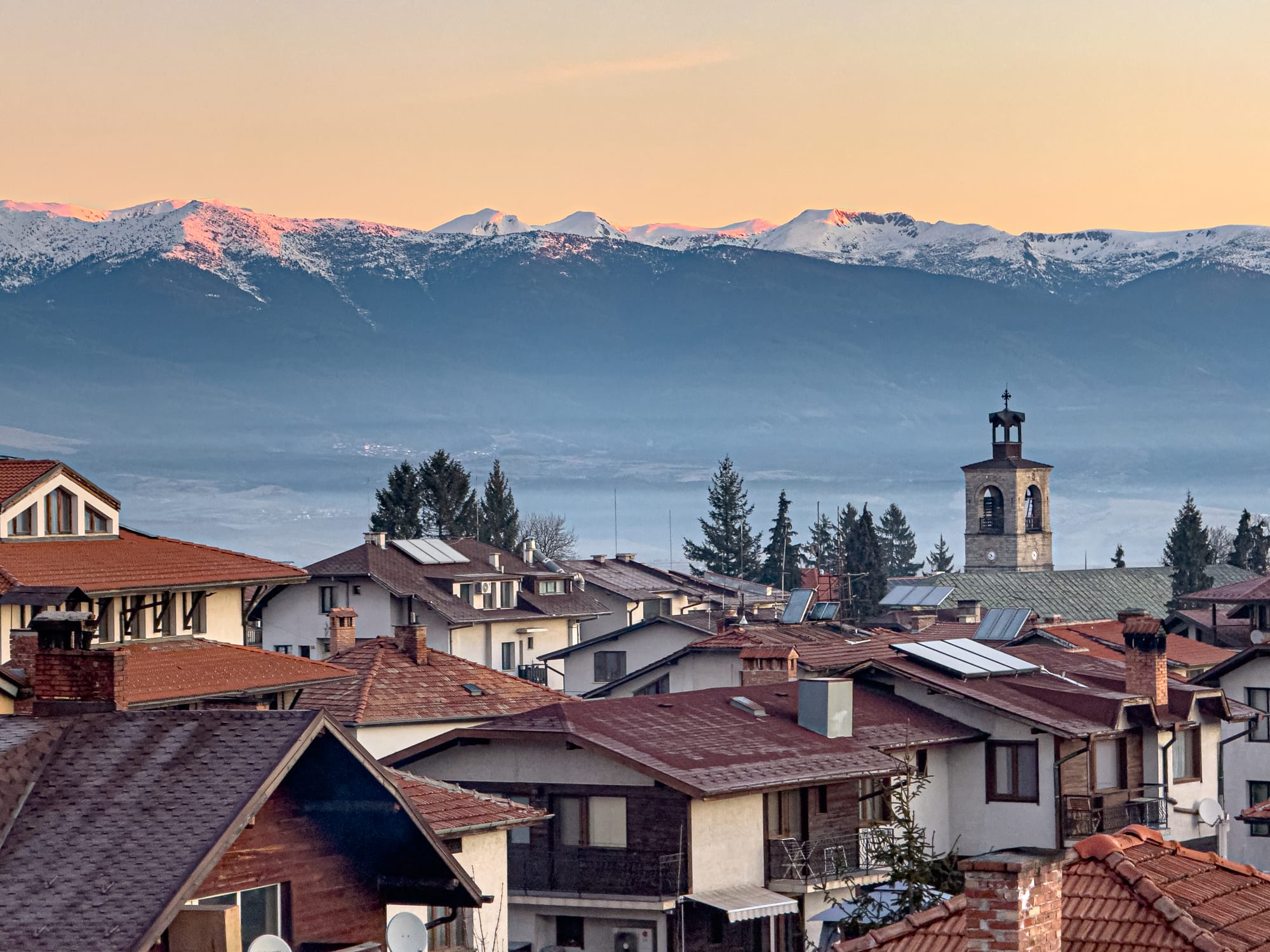 Snow-dusted Pirin Mountains glowing at sunset behind the rooftops and church tower of Bansko, Bulgaria, with warm light casting over the town