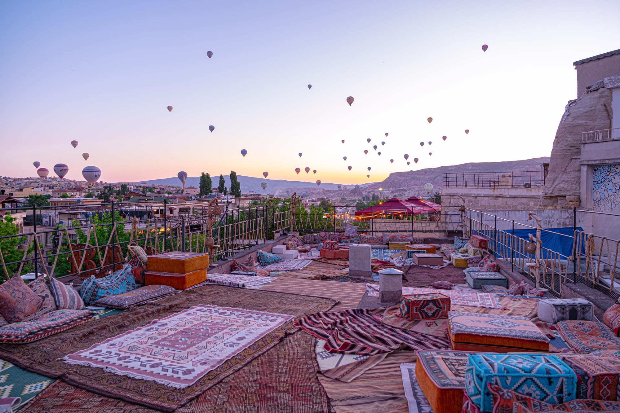 Rooftop terrace in Göreme, Cappadocia, covered with patterned rugs and colorful floor cushions, facing a wide valley with hot air balloons above