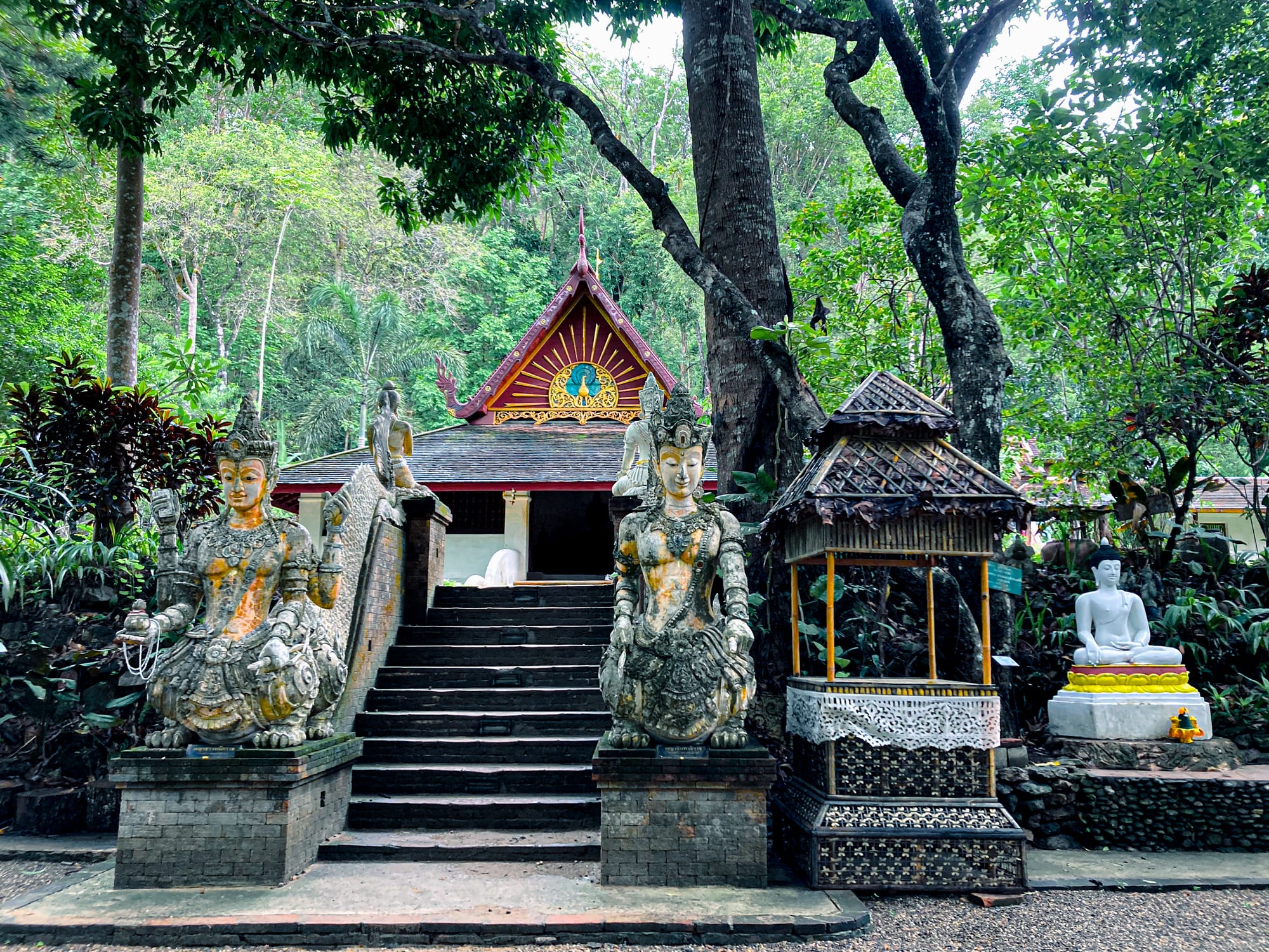 Ornate temple entrance in Chiang Mai with two guardian statues, a small shrine, and a seated white Buddha surrounded by lush green forest