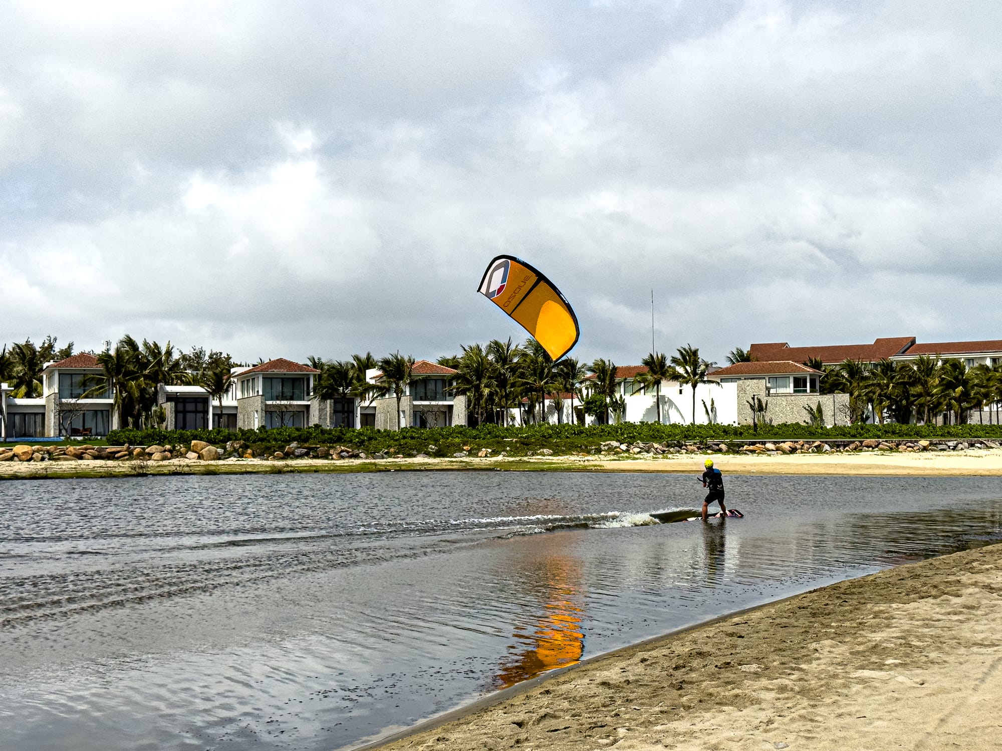 A kitesurfer rides the water near Hội An, holding a bright yellow kite against a cloudy sky, with beachfront villas and palm trees lining the lake