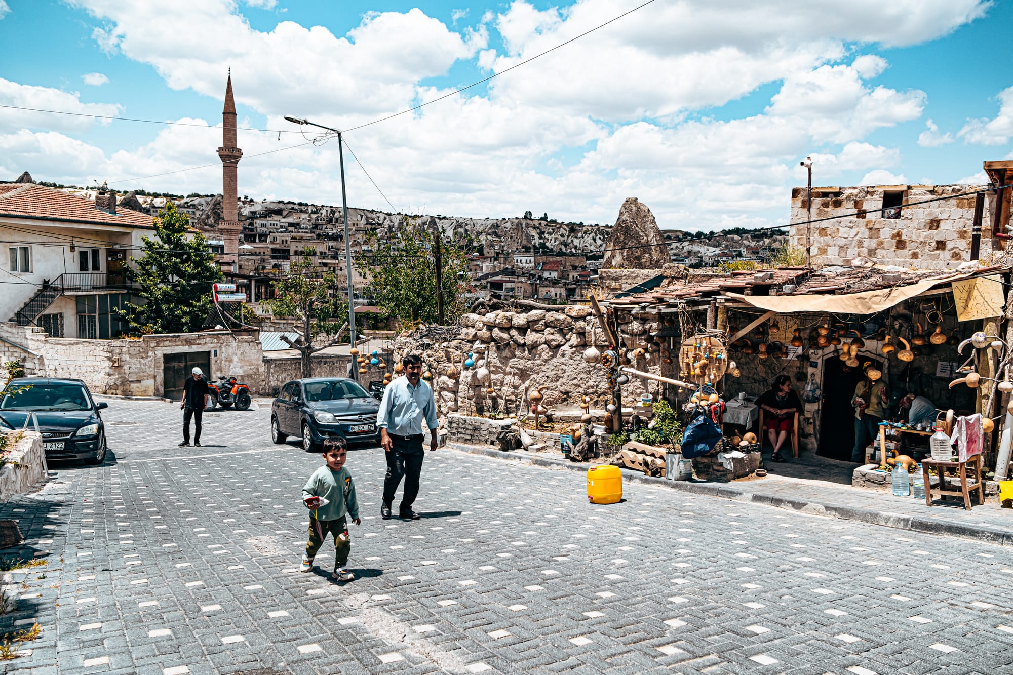 A man and child walk along a cobblestone street in Göreme, passing a rustic shop decorated with hanging gourds and pottery