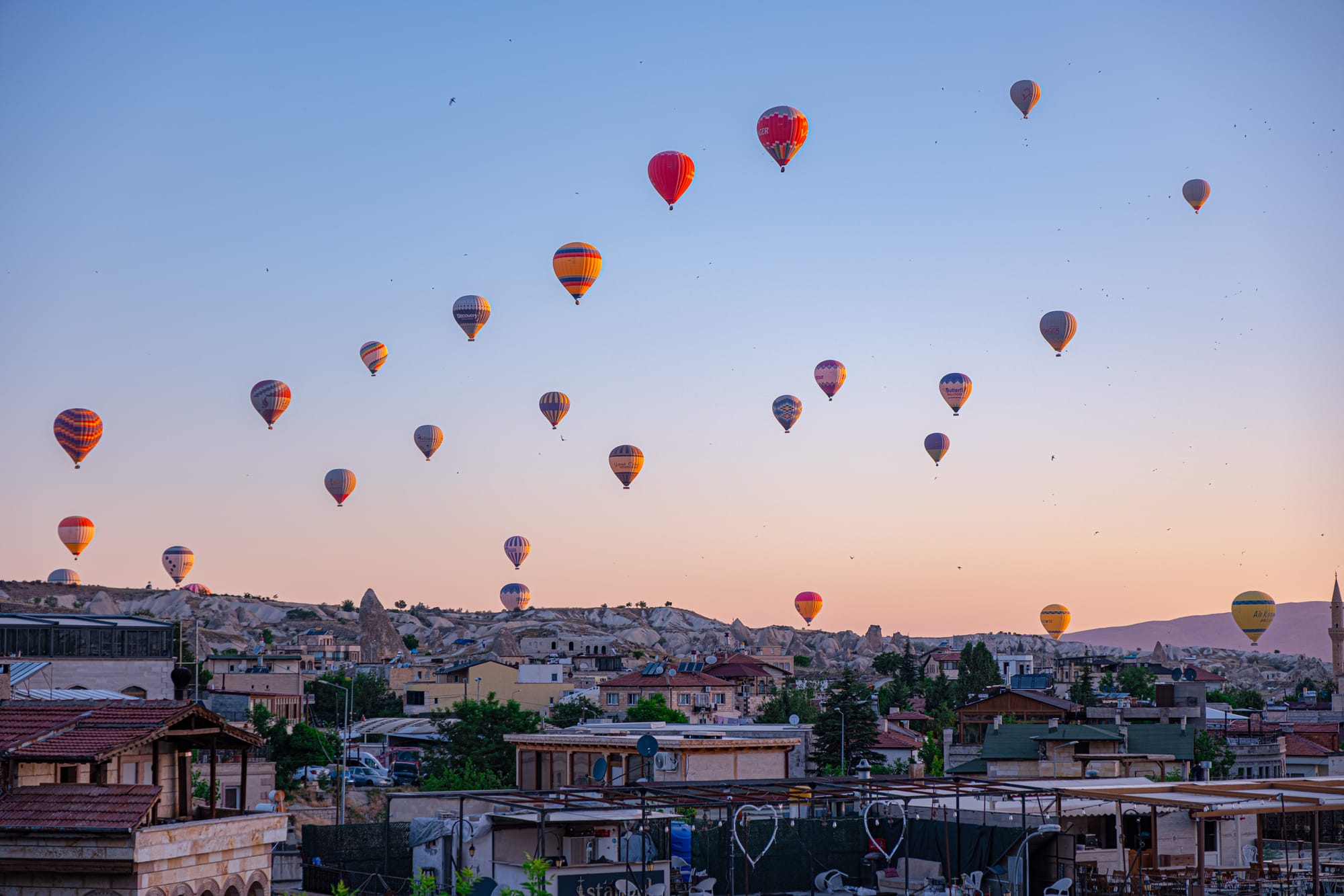Multiple hot air balloons rise above Göreme’s stone houses and fairy chimneys at sunrise, viewed from the rooftop of Nomad Cave Hotel