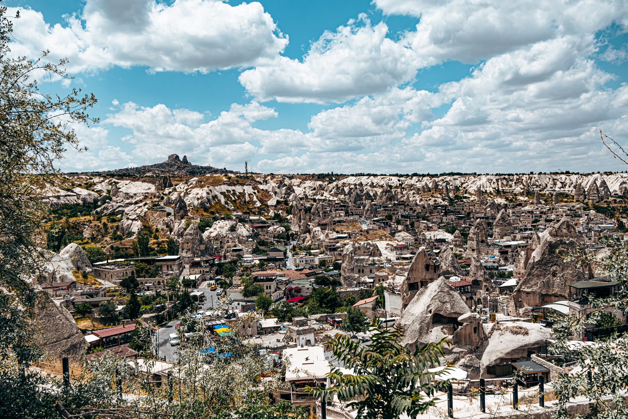 Panoramic photograph of Göreme, Turkey, showing clustered stone buildings, rock-cut dwellings, and surrounding white cliffs under a partly cloudy sky