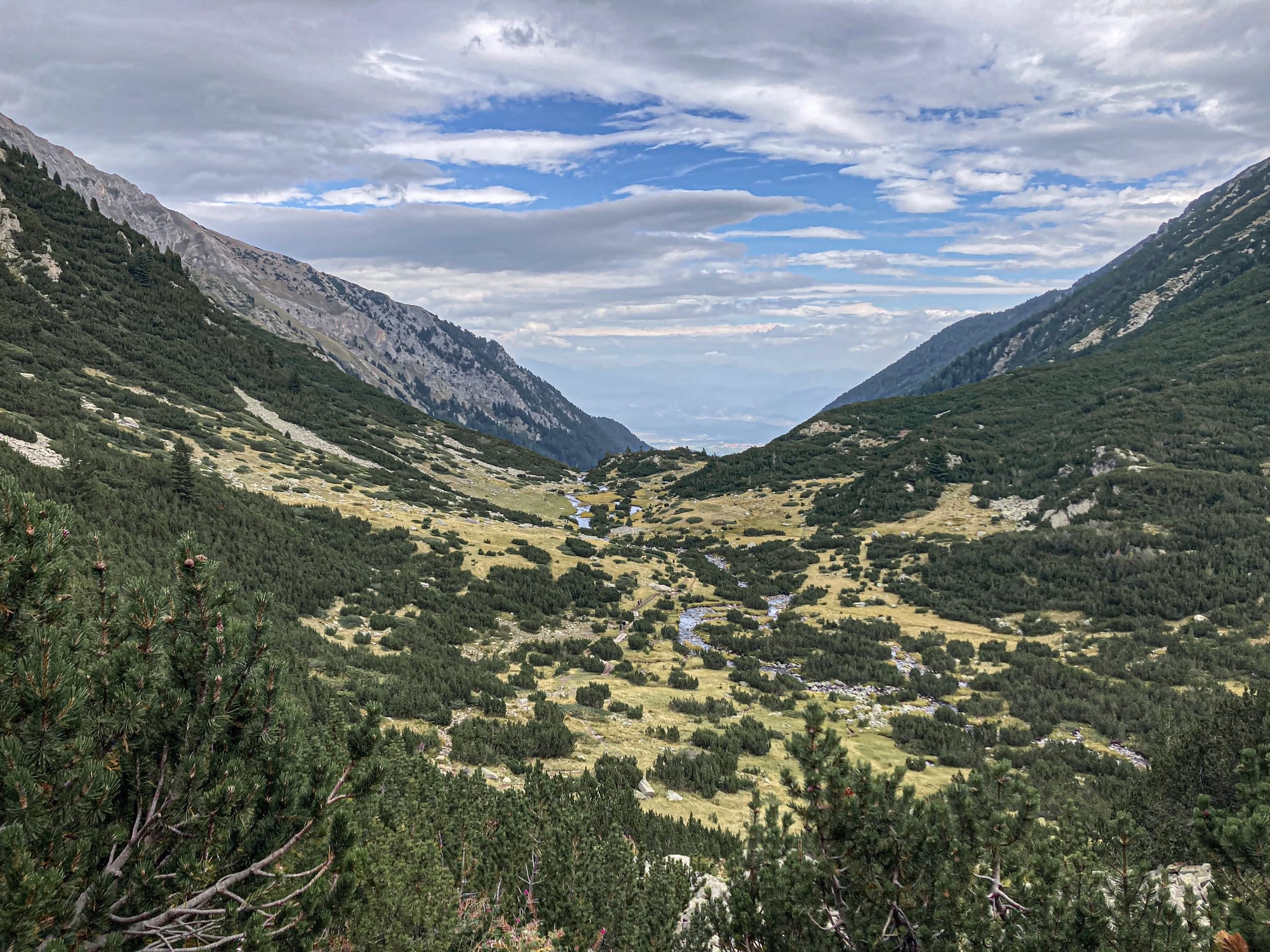 Wide view of a green alpine valley in the Pirin Mountains, with streams cutting through meadows and dark green shrubs, flanked by steep rocky slopes under a cloudy sky with patches of blue