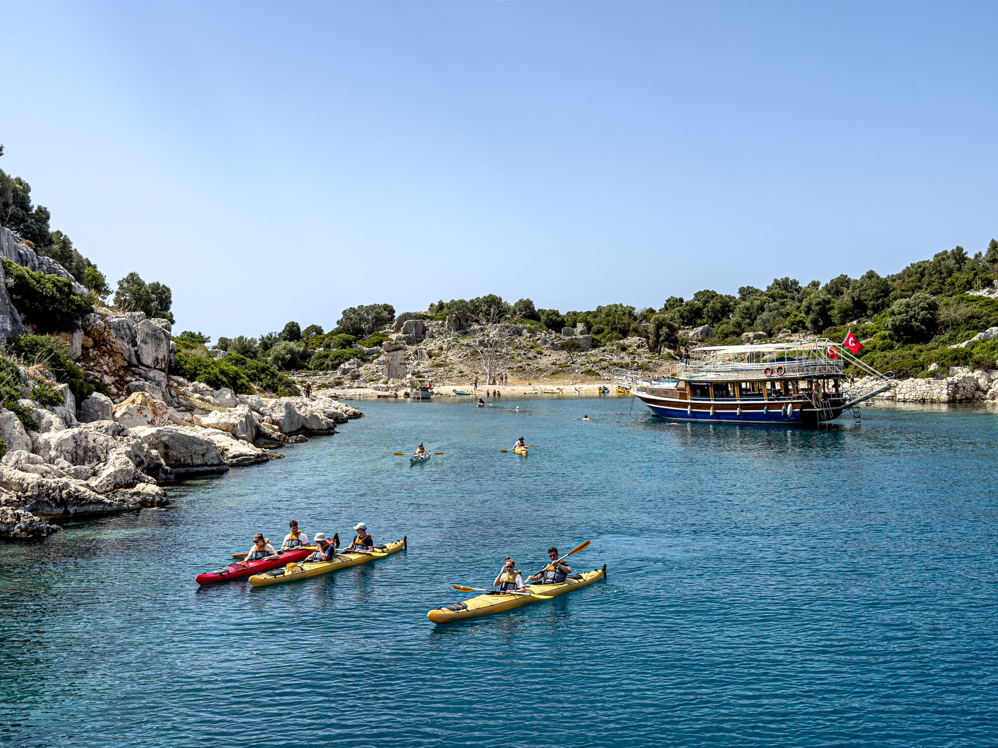 Groups of kayakers paddling through a calm bay near a moored Turkish boat at Kekova, with rocky shores and hillside ruins in the background