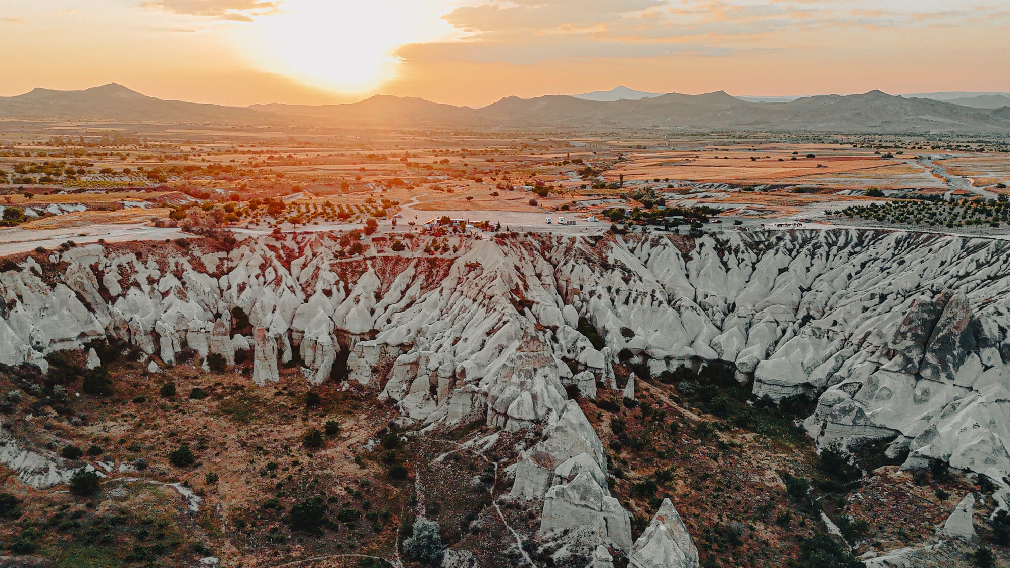 Aerial view of Love Valley in Cappadocia at sunset, with the sun low on the horizon illuminating the pale, pillar-like rock formations in warm golden light, surrounded by expansive fields and distant mountains