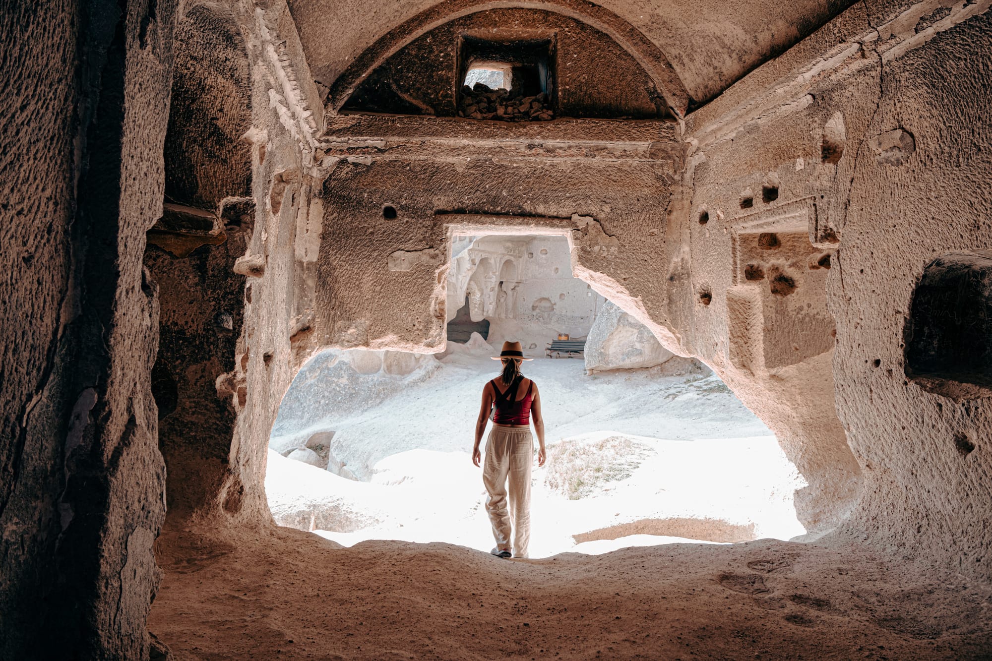 Woman standing inside Selime Monastery cave chamber looking out toward sunlit rock formations and benches