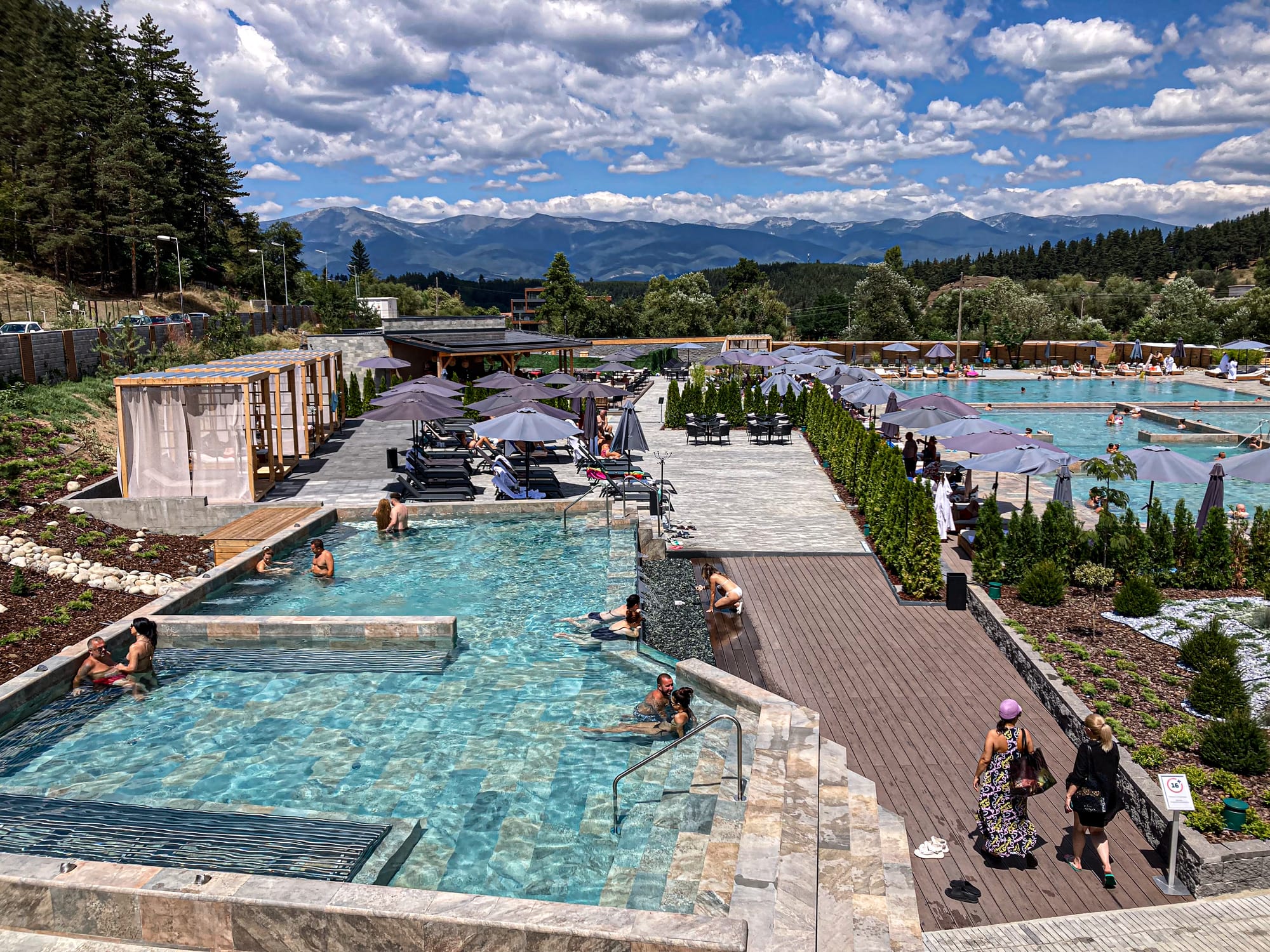 Outdoor mineral pools at Pulse Therme in Banya, Bulgaria, featuring people lounging in thermal waters, shaded seating areas, and mountain scenery in the distance