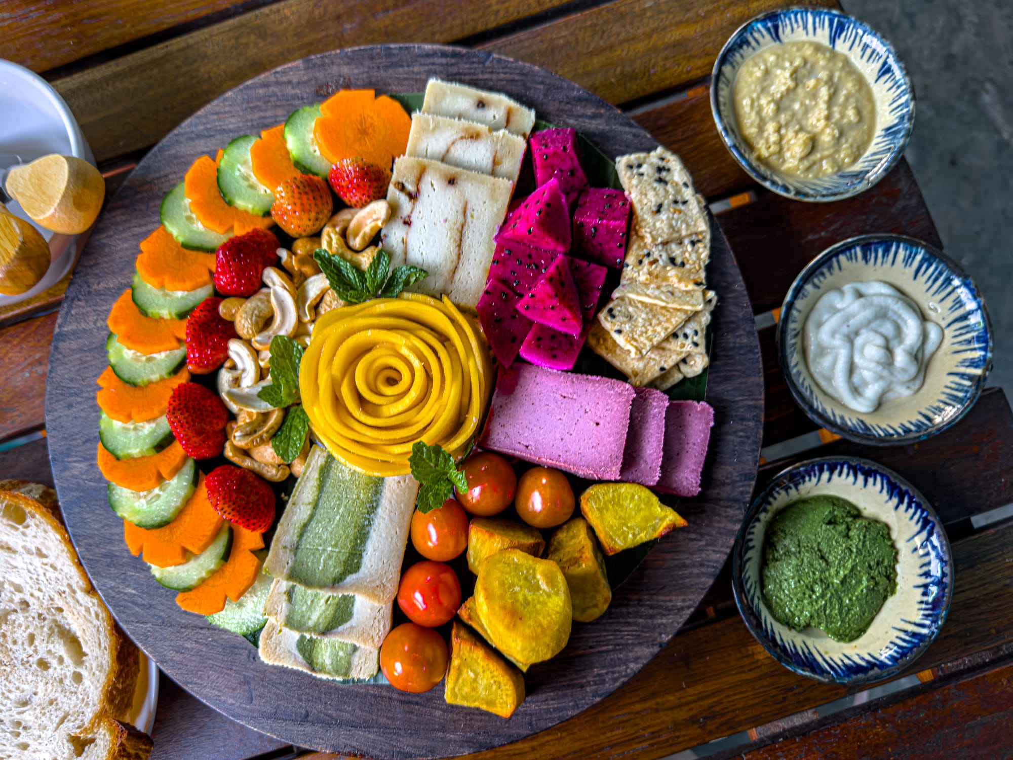 Vegan cheese platter at Chickpea Eatery in Hội An with nut-based cheeses, crackers, fresh strawberries, dragon fruit, cucumber, carrot slices, cherry tomatoes, cashews, roasted sweet potato, and a carved mango rose centerpiece, served with three dipping sauces