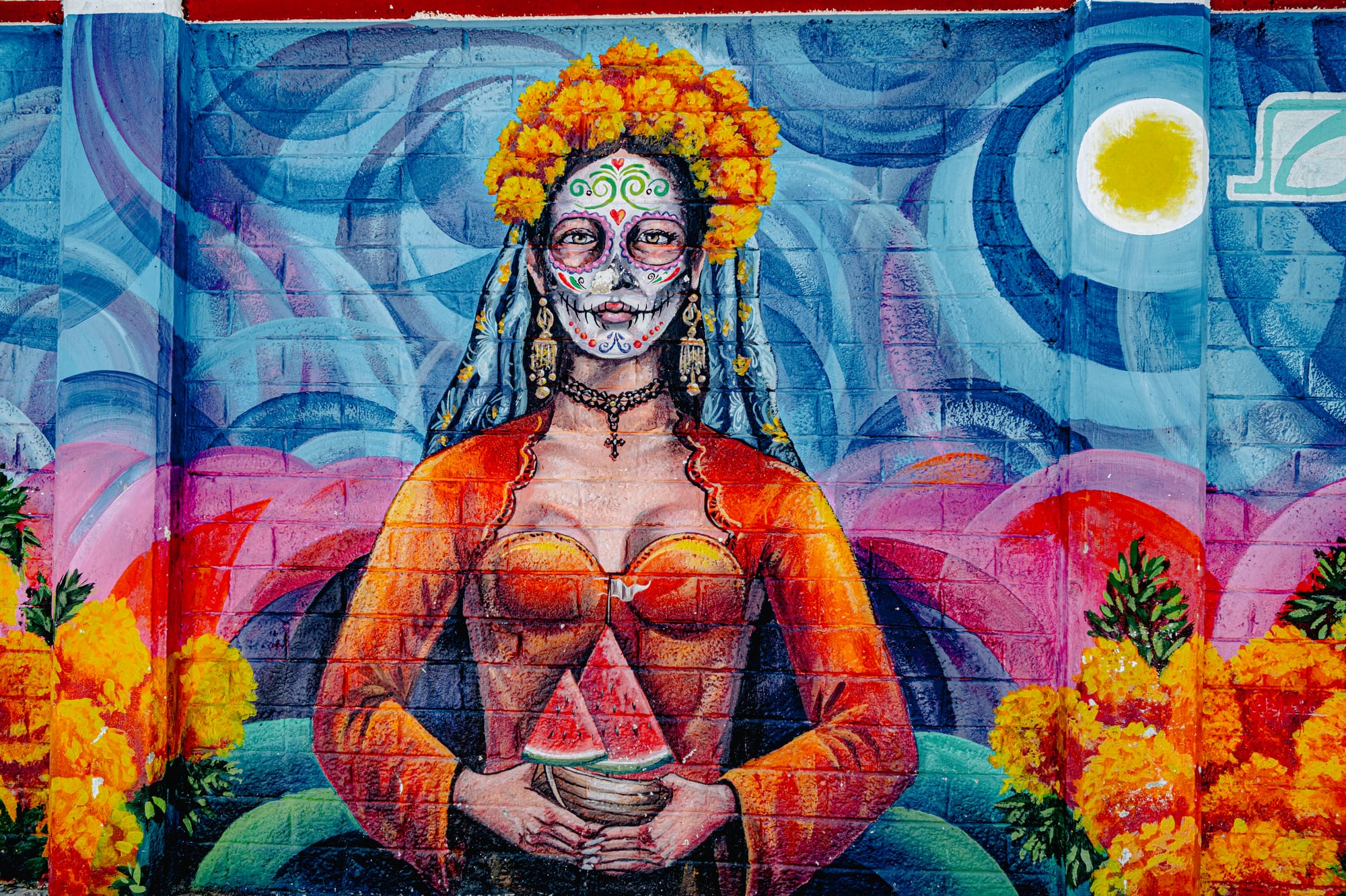 A mural in San Cristóbal de las Casas portraying La Catrina with a painted skull face, marigold crown, and vibrant orange dress holding slices of watermelon, symbolizing vitality, remembrance, and the blending of life and death in Day of the Dead traditions