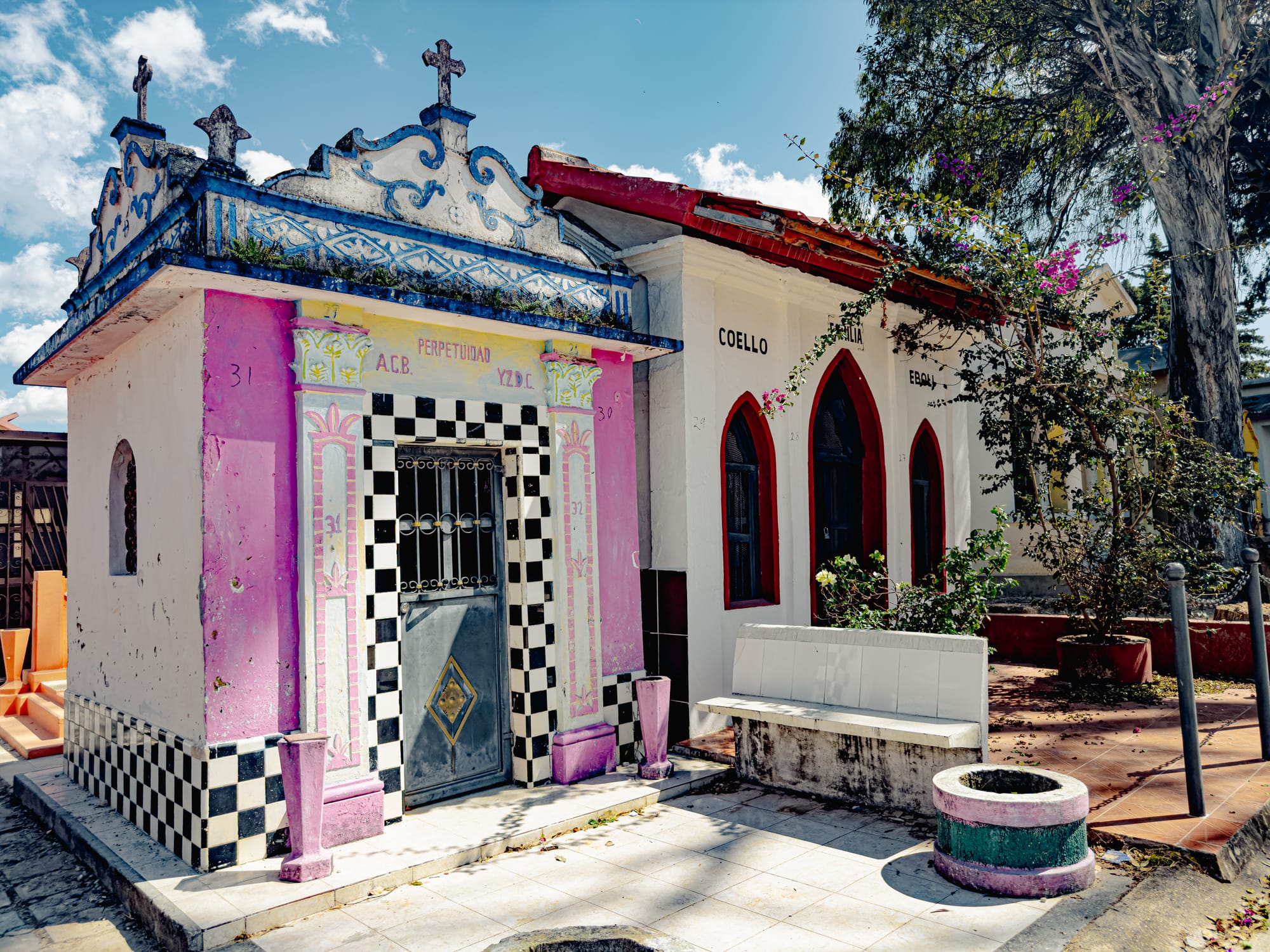 A pair of ornate mausoleums in the Municipal Pantheon of San Cristóbal de las Casas—one painted in pastel pink, black, and white tiles, and the other in cream and red—showcasing Chiapas’ mix of folk art and reverence in Day of the Dead traditions