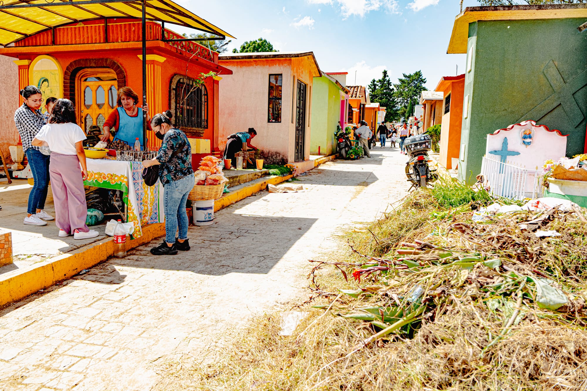 A lively scene inside the Municipal Pantheon of San Cristóbal de las Casas, where women gather around a food stand beside colorful tombs, chatting and eating as families clean and decorate graves ahead of Día de los Muertos