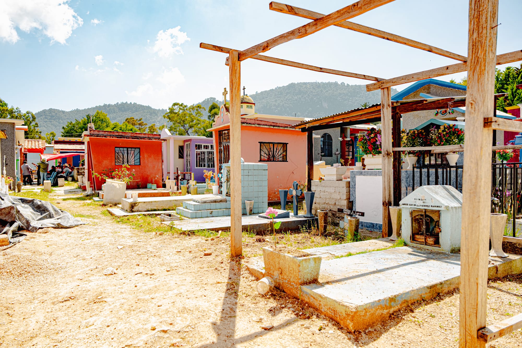 View of the Municipal Pantheon in San Cristóbal de las Casas, showing colorful tombs, flowers, and small mausoleums beneath wooden frames and bright sunlight, with families working in the background to clean and decorate for Day of the Dead
