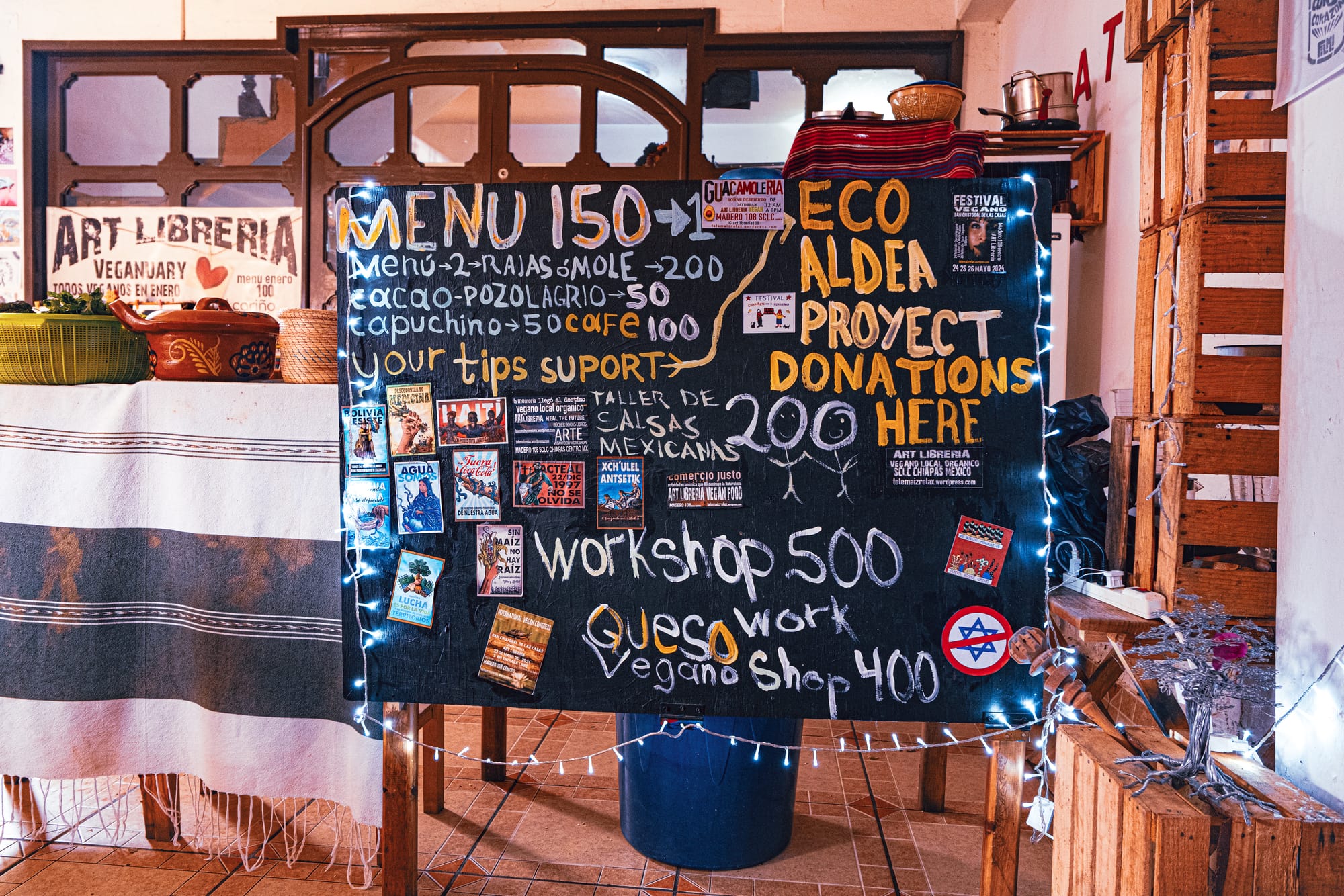 A hand-painted chalkboard at Art Libreria in San Cristóbal de las Casas showing vegan menu prices, workshop listings, and Eco Aldea donation information, with baskets, clay pots, and wooden shelves around it