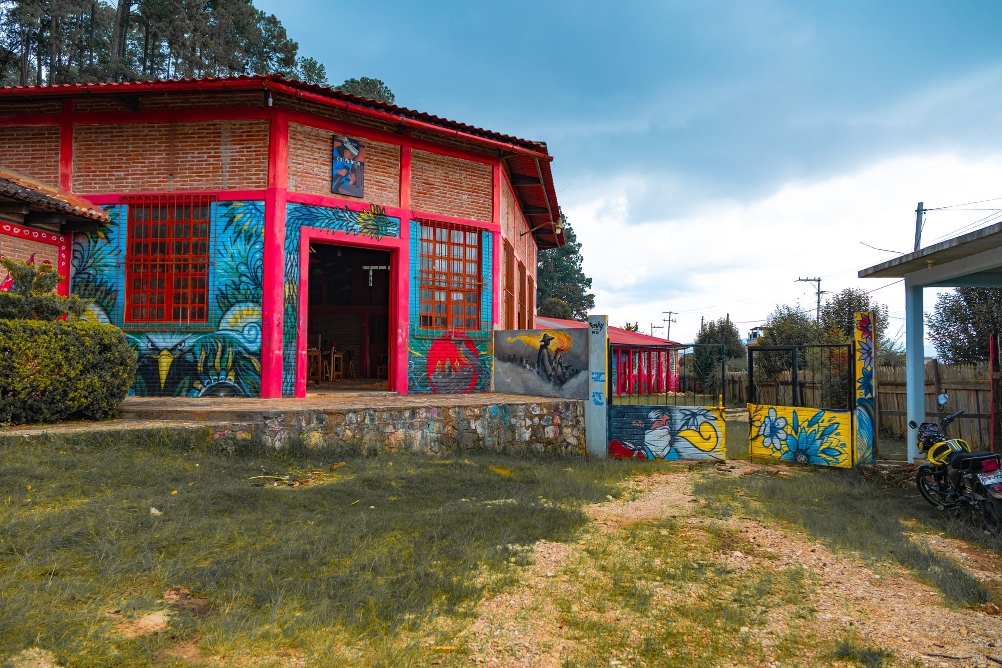Colorful mural-covered building at CIDECI-Unitierra in Chiapas, featuring bright red trim, indigenous-inspired artwork, and an open doorway leading into a communal learning space surrounded by trees and rustic pathways