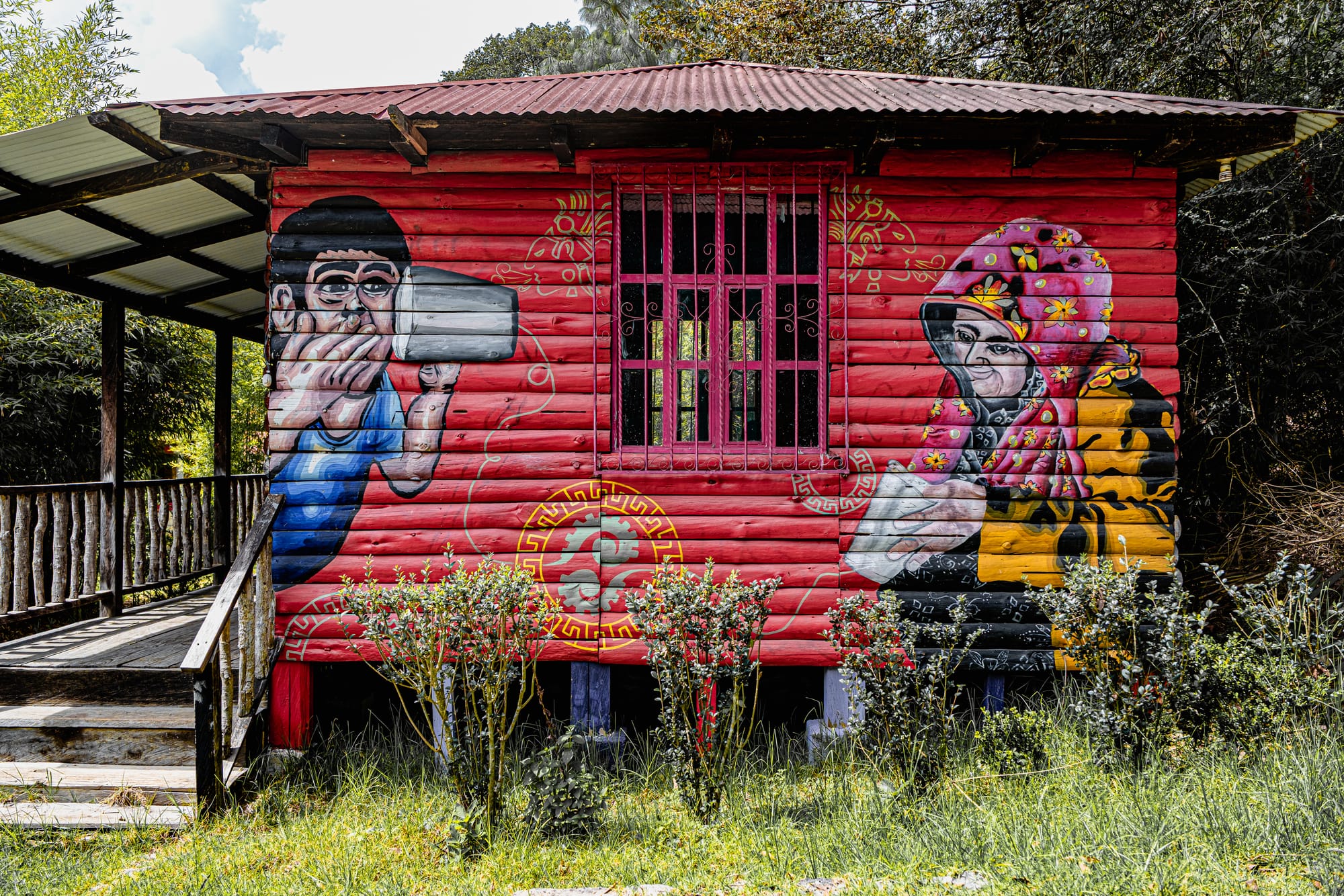 Mural on a red wooden building at CIDECI depicting two stylized figures drinking from cups, with vibrant floral patterns and decorative motifs surrounding a pink-barred window, set beside a rustic walkway and greenery