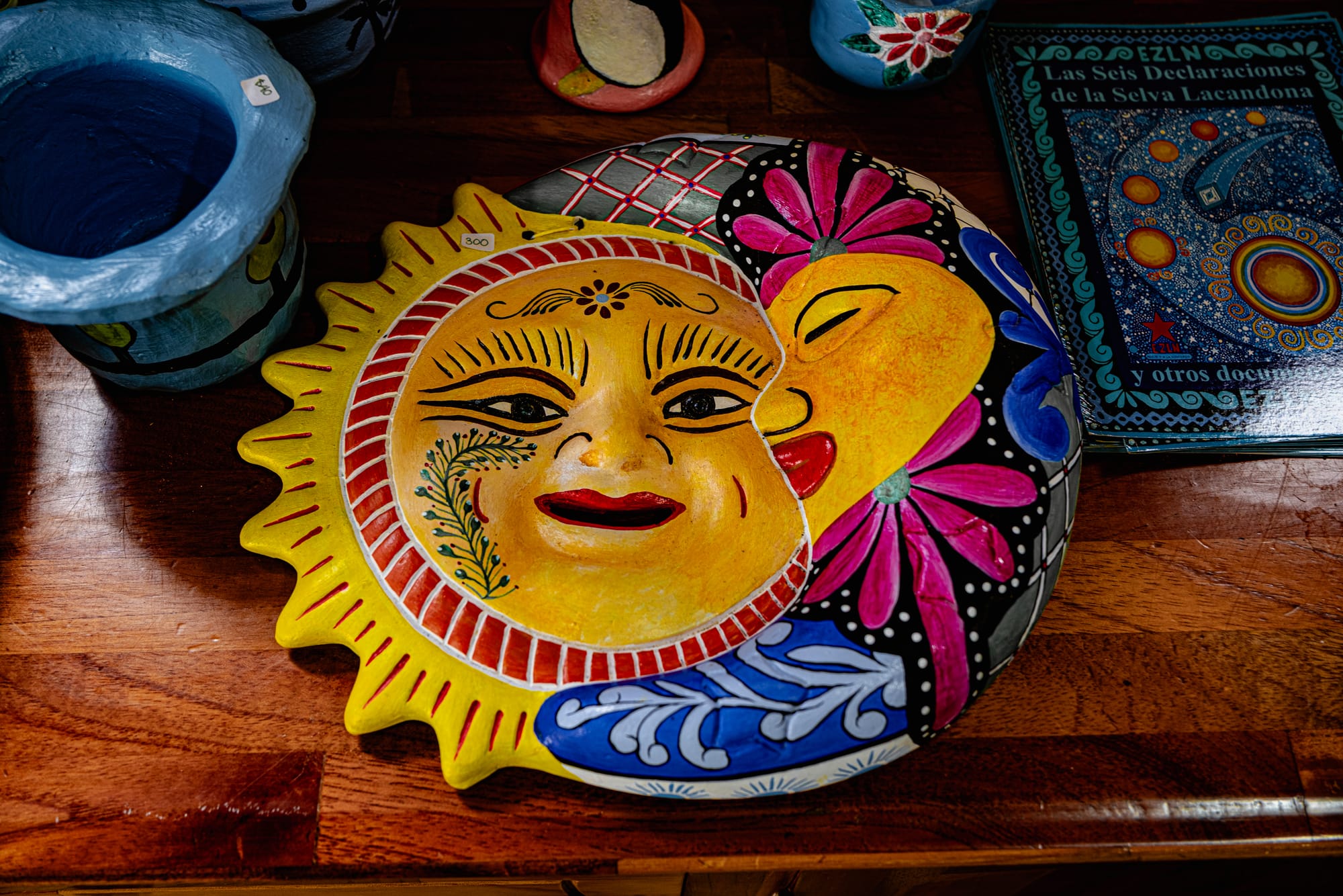 Hand-painted ceramic sun and moon decoration displayed on a wooden table at CIDECI, surrounded by colorful pottery and a book titled ‘Las Seis Declaraciones de la Selva Lacandona'