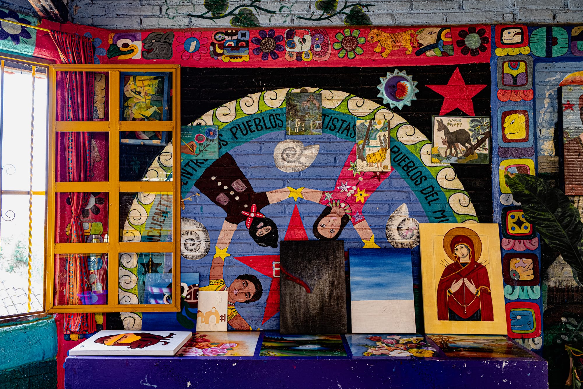 Mural inside a colorful room at CIDECI featuring masked figures holding hands around a red star, surrounded by flowers, animals, and Mayan-inspired designs, with canvases and paintings displayed along the bottom
