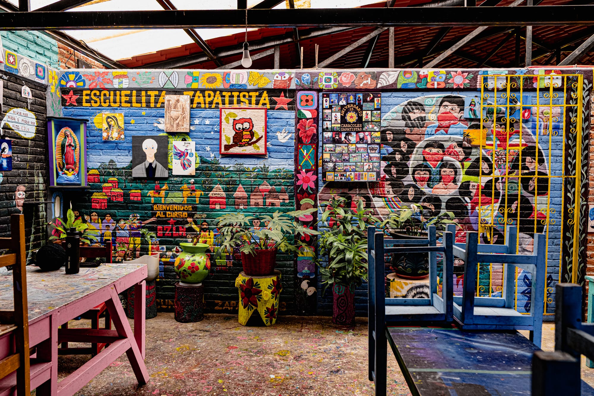 Colorful mural-filled classroom at CIDECI labeled ‘Escuelita Zapatista,’ featuring painted walls with Zapatista figures, village scenes, symbols, and student artwork, alongside brightly painted tables, chairs, and potted plants