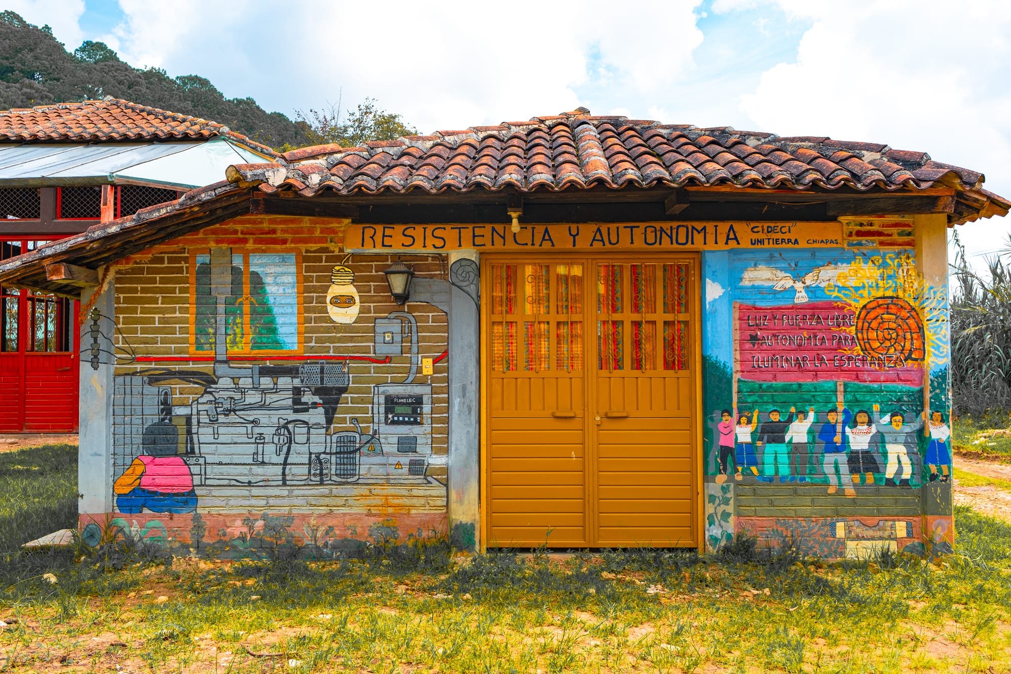 Mural-covered building at CIDECI in Chiapas with the words ‘Resistencia y Autonomía,’ featuring painted scenes of community power generation, collective organizing, and the message ‘Luz y fuerza libre y autonomía para iluminar la esperanza,’ under a tiled roof with mountains in the background