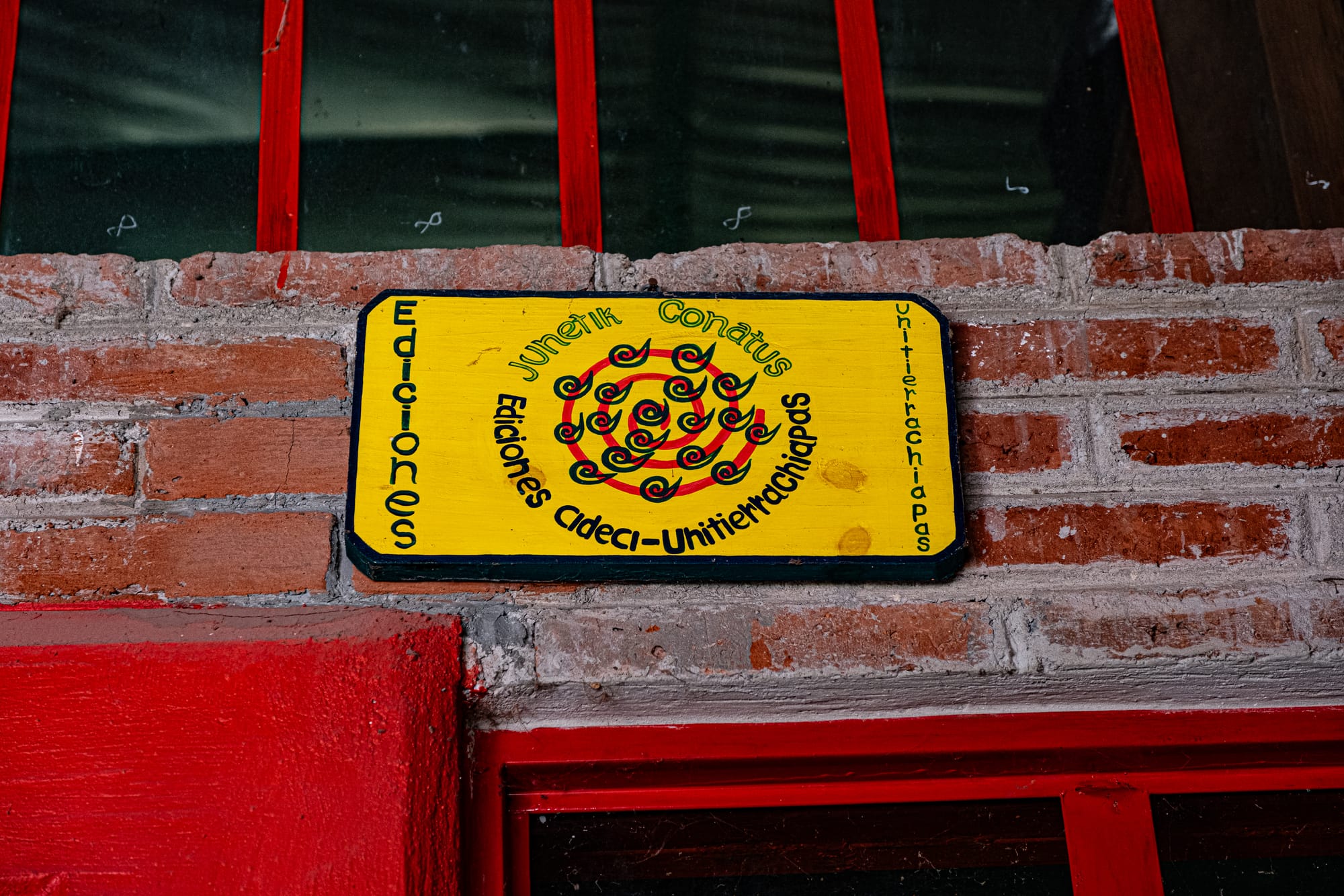 Yellow wooden sign for Ediciones CIDECI-Unitierra Chiapas mounted on a brick wall, featuring a spiral red-and-black symbol and decorative lettering at an autonomous learning center in San Cristóbal de las Casas