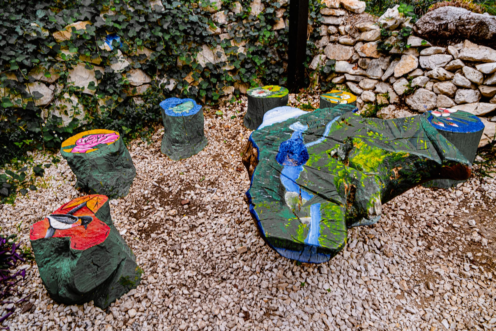 Painted tree-stump seats and a large carved table in an outdoor garden area at CIDECI-Unitierra, decorated with colorful nature-inspired art and surrounded by stone walls and ivy
