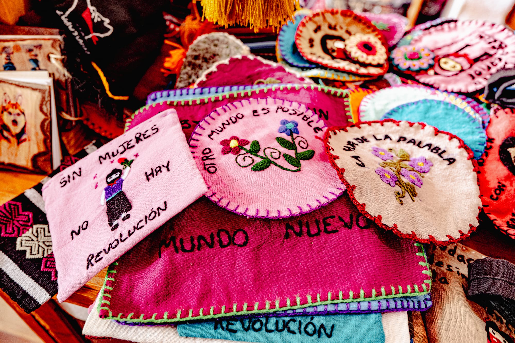 Colorful handmade Zapatista embroidery patches with feminist and revolutionary messages, including stitched phrases like ‘Sin mujeres no hay revolución’ and ‘Otro mundo es posible,’ displayed on a table at an artisan market in Chiapas