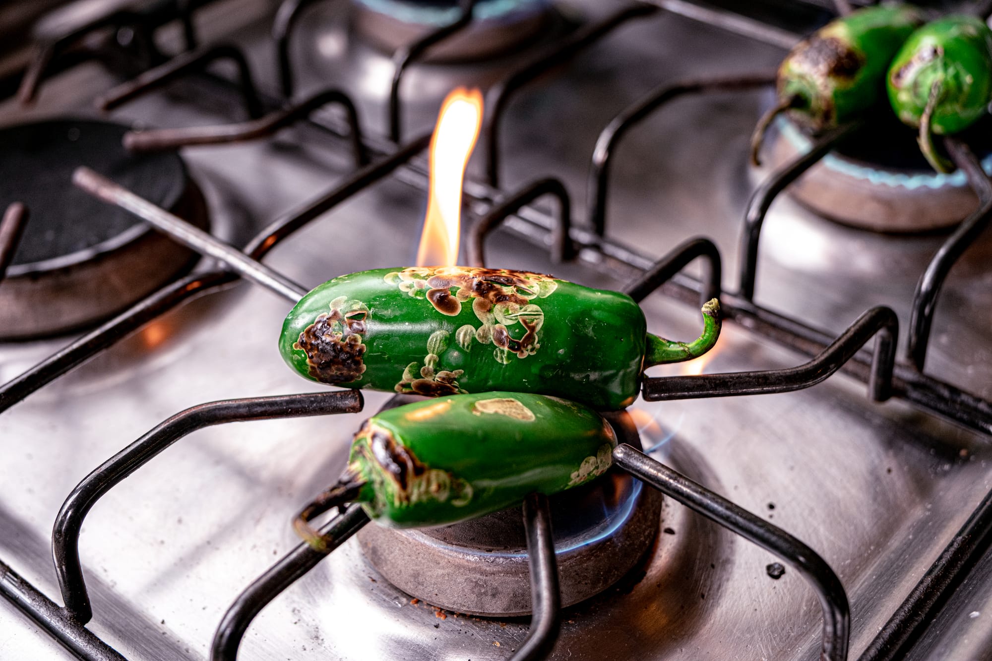 Two green jalapeños blistering and charring directly over a gas flame on Itzel’s stovetop