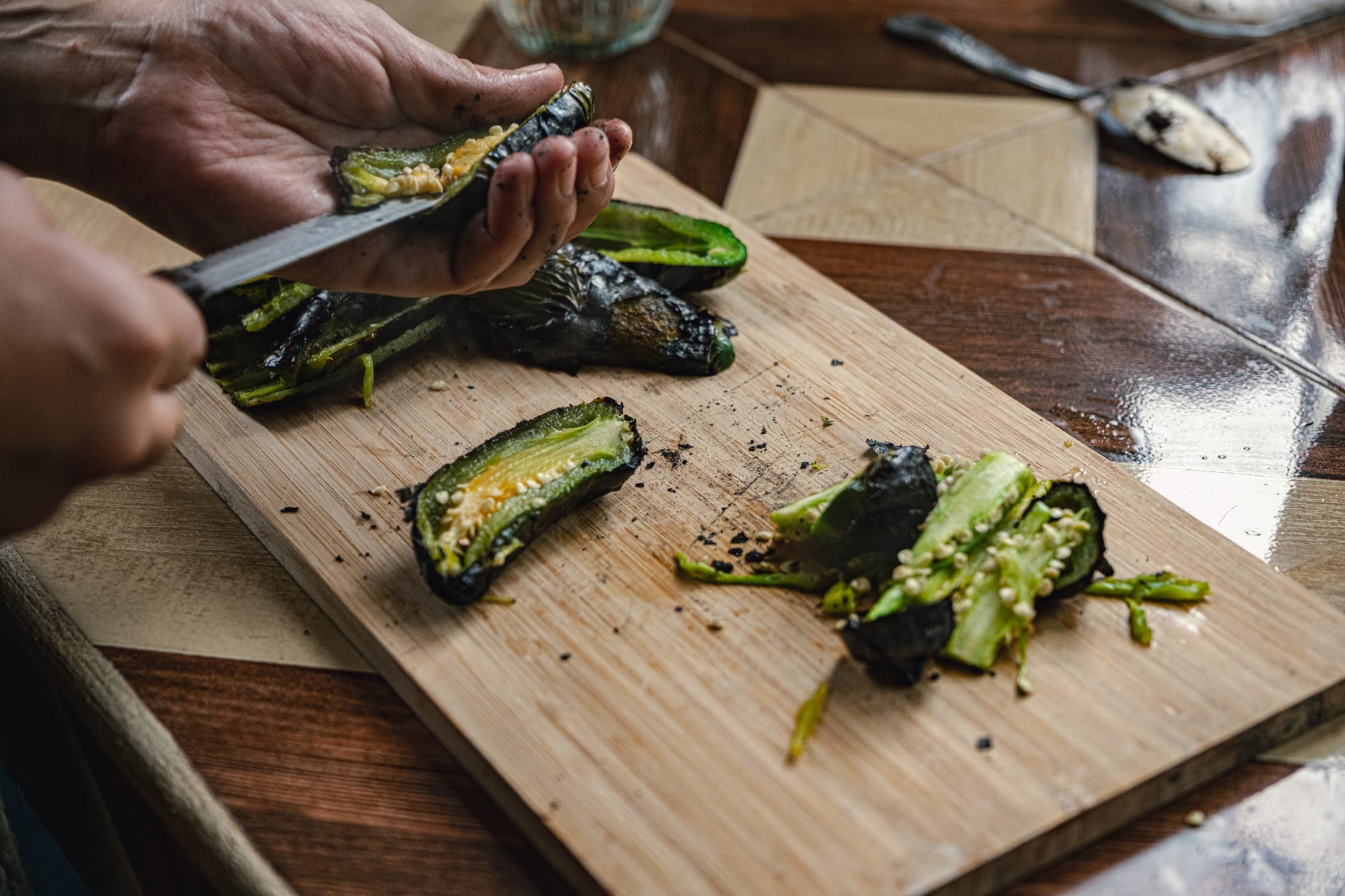 Hands slicing open fire-roasted jalapeños on a wooden cutting board, the charred skins and seeds scattered across the surface