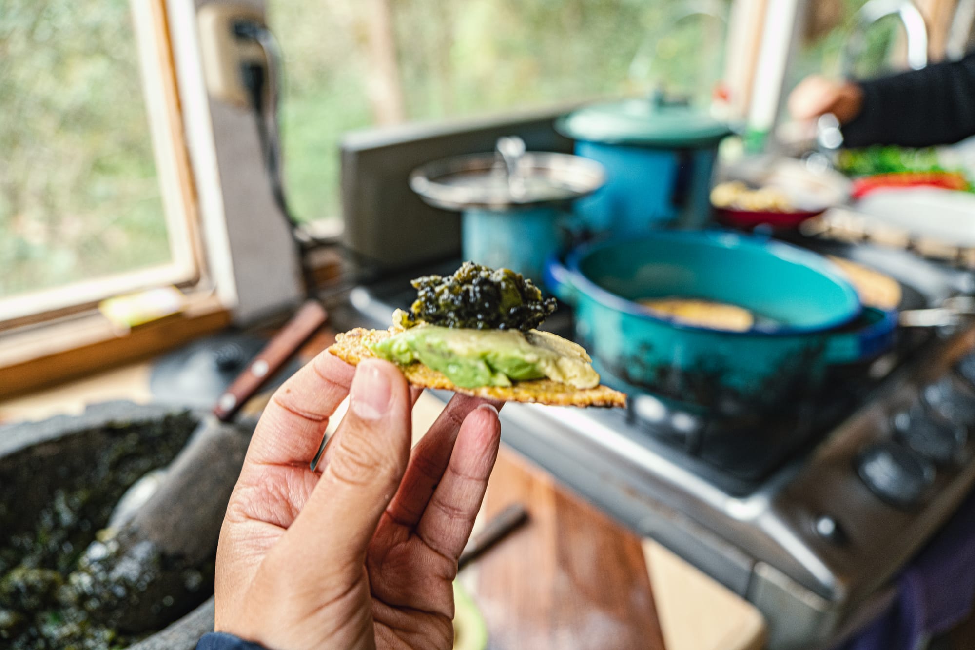 A hand holding a small crisp topped with guacamole and charred jalapeño salsa, with pots and pans in the background of Itzel’s kitchen
