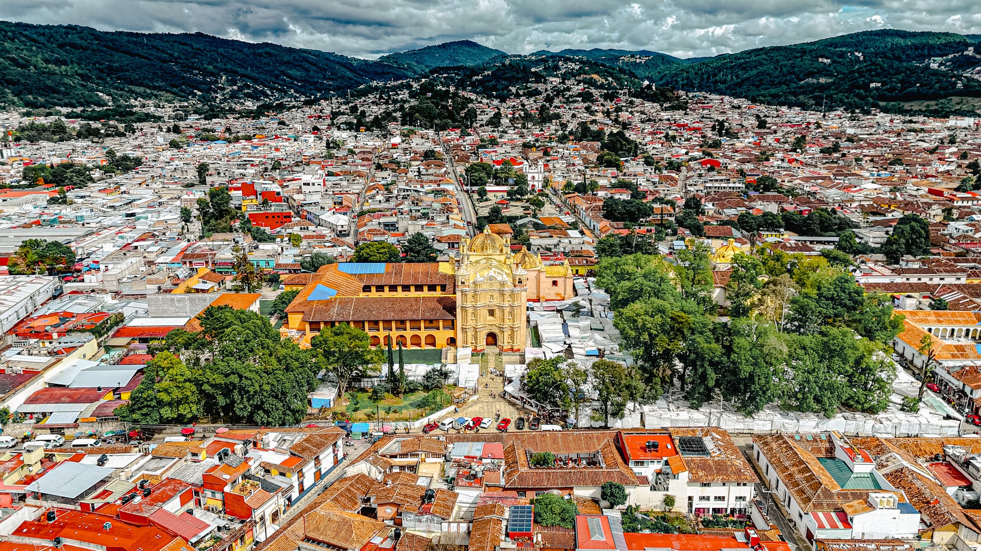 Aerial view of San Cristóbal de las Casas showing Templo de Santo Domingo de Guzmán, its adjacent former convent, surrounding rooftops, and forested hills in Chiapas, Mexico