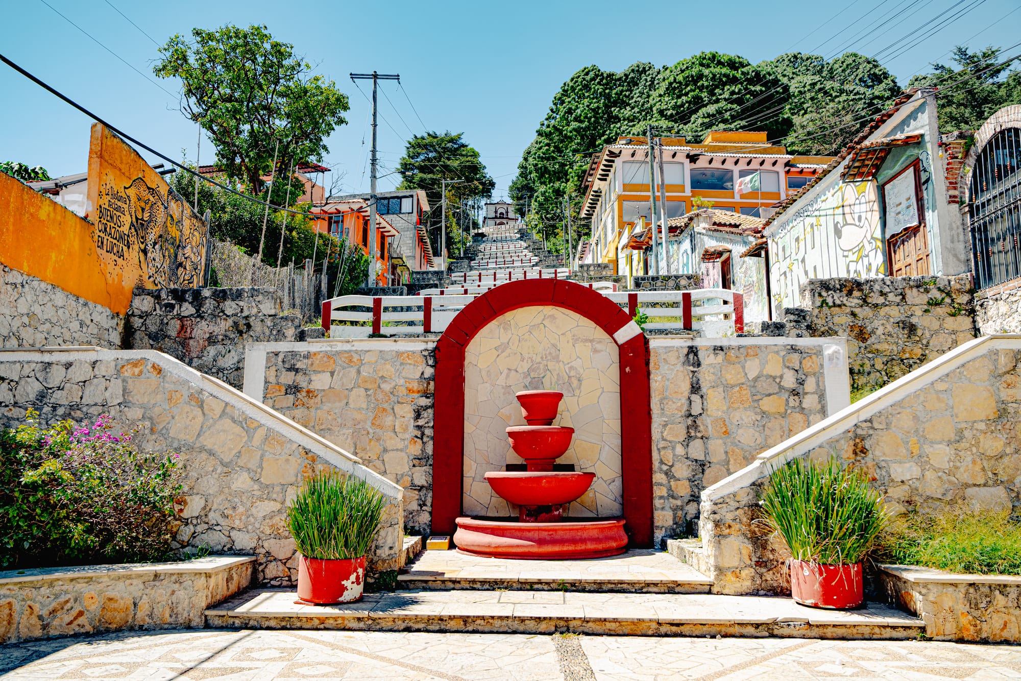 Fountain and landing along the zigzagging staircase to Iglesia de San Cristóbalito in San Cristóbal de las Casas, Chiapas, Mexico, surrounded by stone walls, street art murals, and hillside neighborhood houses