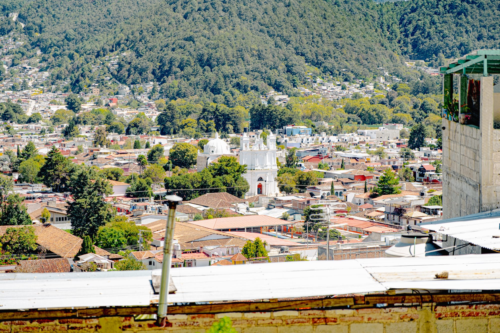 View over San Cristóbal de las Casas from the hillside near Iglesia de San Cristóbalito, showing city rooftops, surrounding mountains, and the white façade of a church below in Chiapas, Mexico