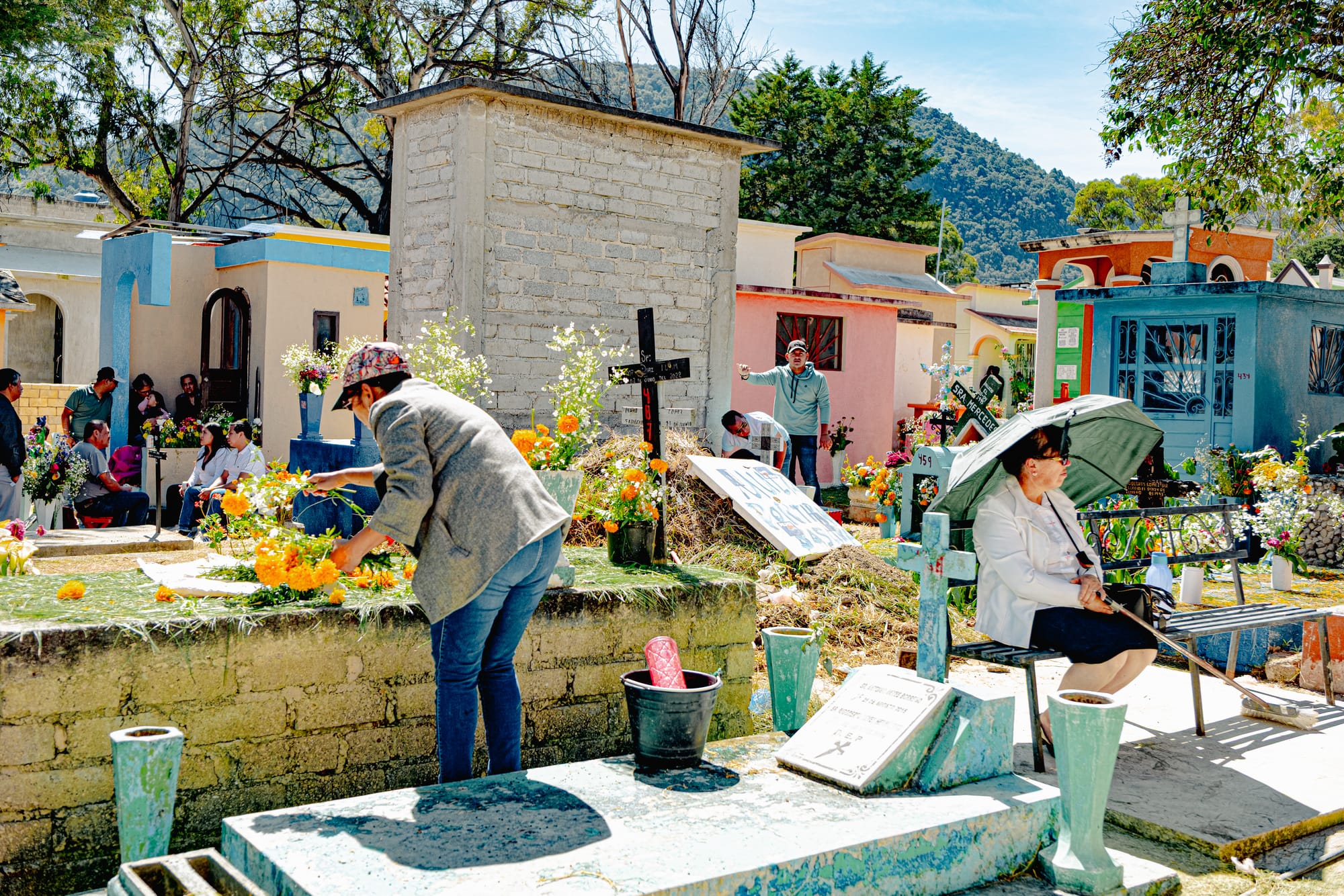 Día de Muertos at San Cristóbal Municipal Pantheon in San Cristóbal de las Casas, Chiapas, Mexico, families decorating above ground tombs with marigolds and flowers, people sitting beside graves under umbrellas, colorful house like mausoleums and community gathering inside the city cemetery