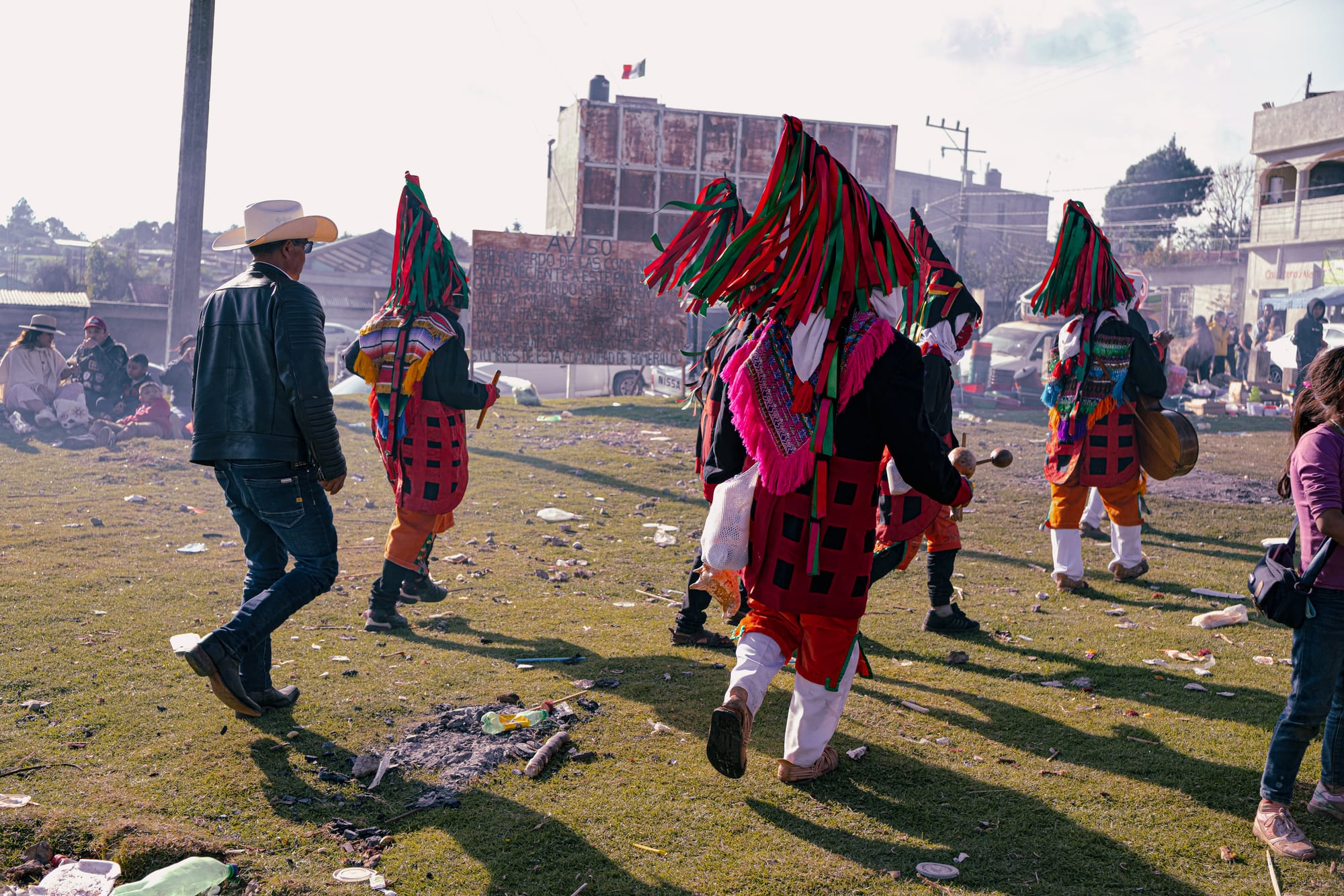 Día de Muertos at Romerillo Cemetery Chiapas, indigenous musicians in traditional ribbon headdresses and woven ceremonial clothing walking from the hilltop cemetery toward the surrounding fairgrounds, live music procession connecting graves and festival space, pine covered tombs and marigold offerings behind them, local Day of the Dead celebration near San Cristóbal de las Casas