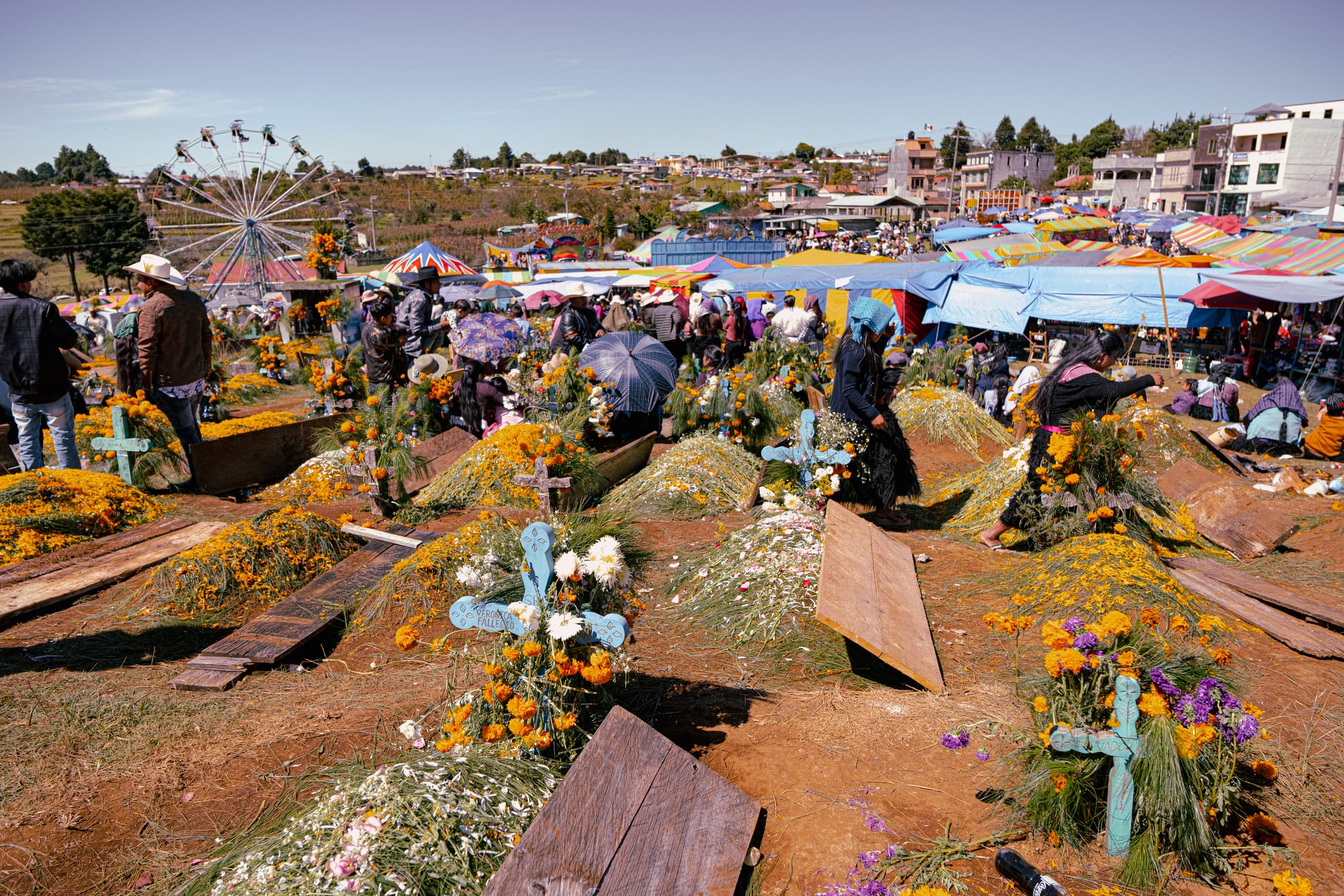 Día de Muertos at Romerillo Cemetery Chiapas, hillside cemetery covered in pine needles and marigolds with wooden crosses, families visiting graves during Day of the Dead celebrations, crowded local cemetery with festival tents and amusement park visible nearby, Romerillo Cemetery near San Cristóbal de las Casas traditional indigenous Day of the Dead scene