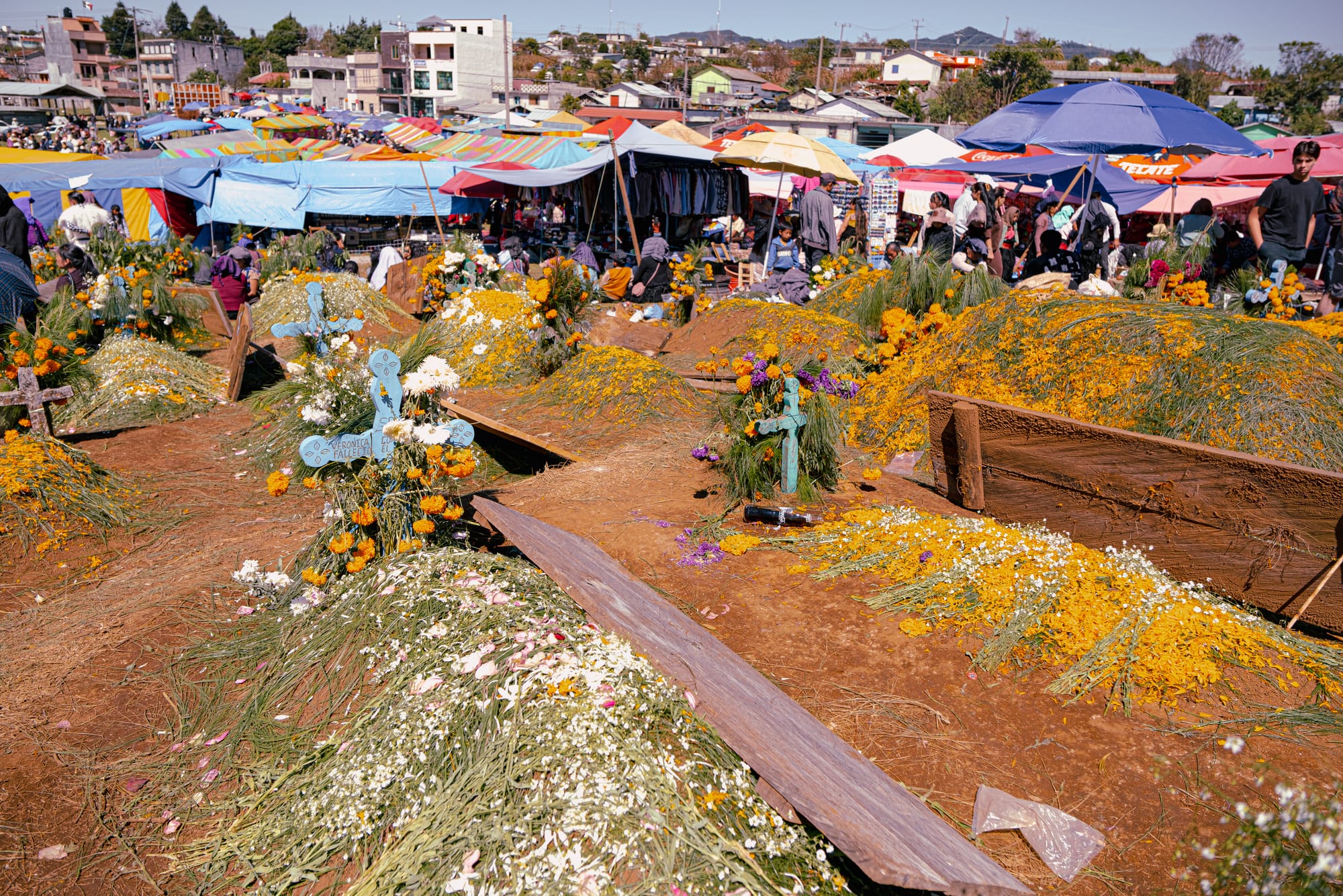 Día de Muertos at Romerillo Cemetery Chiapas, densely packed hilltop cemetery with pine covered graves and marigold decorated mounds, wooden crosses marking tombs across uneven ground, crowded indigenous Day of the Dead celebration with market tents and vendors visible beside the cemetery, Romerillo Cemetery near San Cristóbal de las Casas festival atmosphere