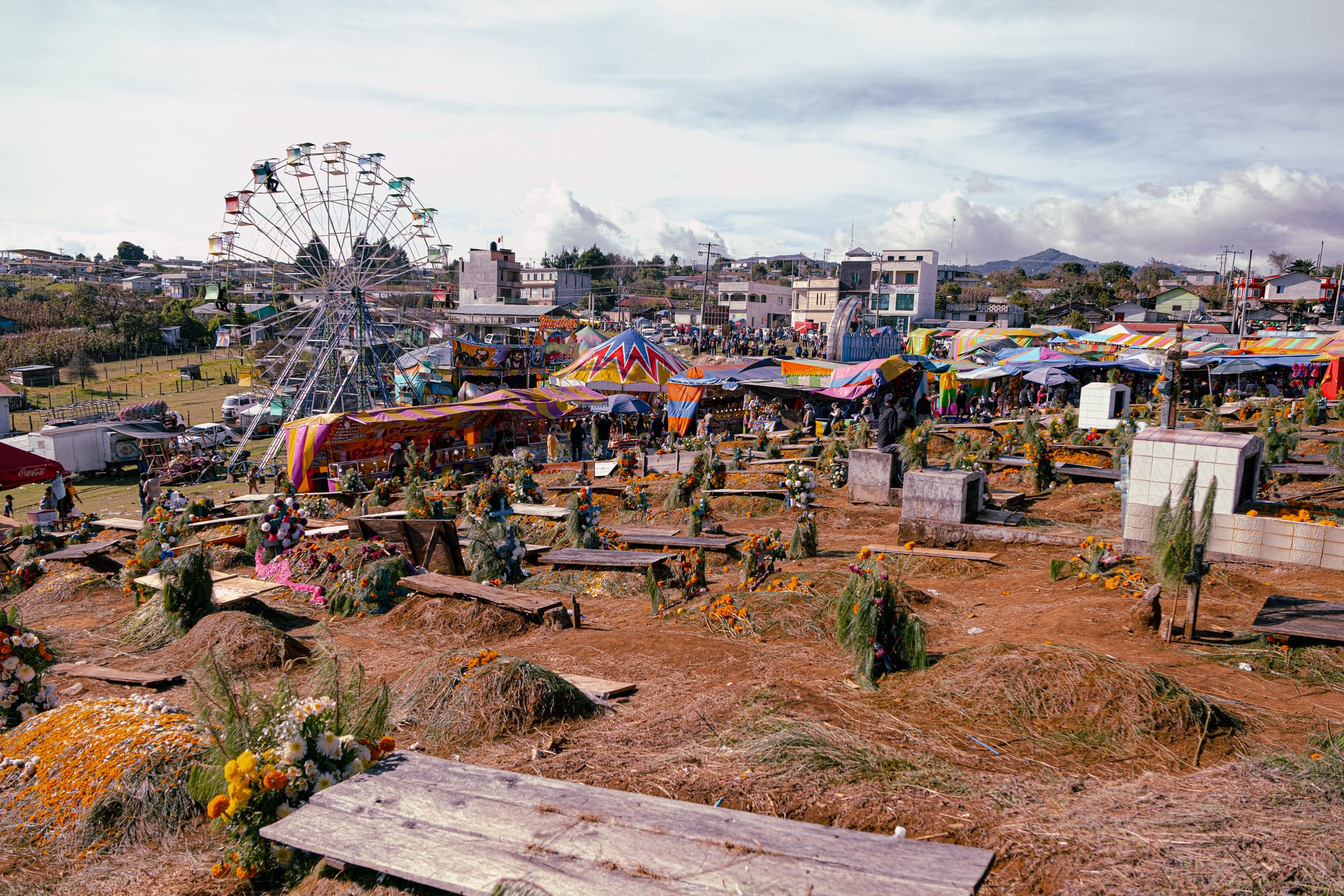 Día de Muertos at Romerillo Cemetery Chiapas, wide panoramic view of hilltop cemetery covered with pine needles and marigold decorated graves, families gathered among wooden crosses and shallow tombs, amusement park rides and colorful festival tents visible beside the cemetery, local indigenous Day of the Dead celebration near San Cristóbal de las Casas