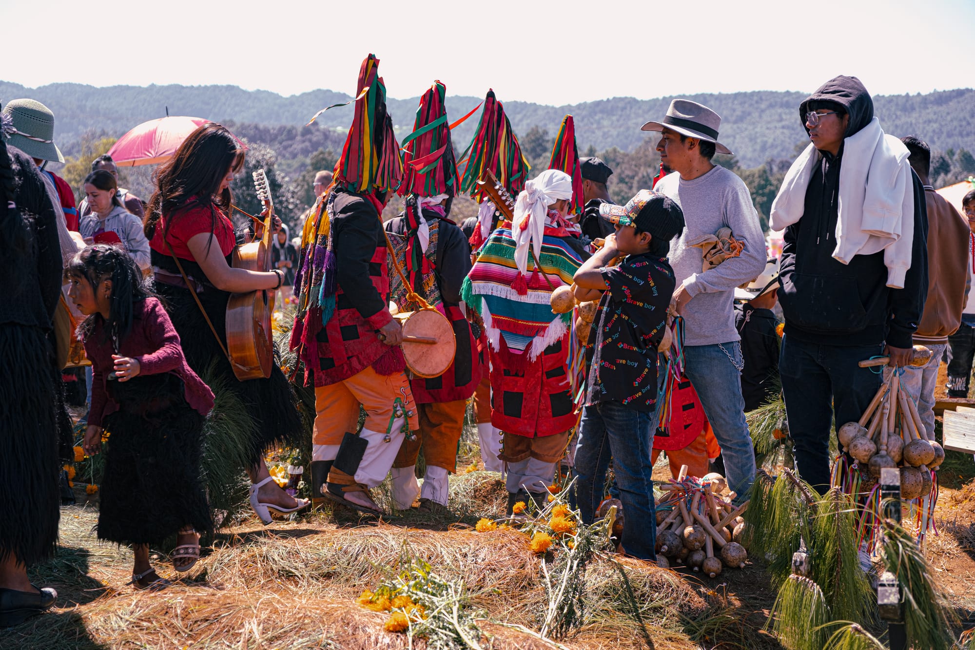 Día de Muertos at Romerillo Cemetery Chiapas, indigenous musicians and families gathered around pine covered graves, traditional ribbon headdresses and woven clothing worn during Day of the Dead, live music with guitar drums and rattles, marigolds and food offerings placed on hilltop graves near San Cristóbal de las Casas