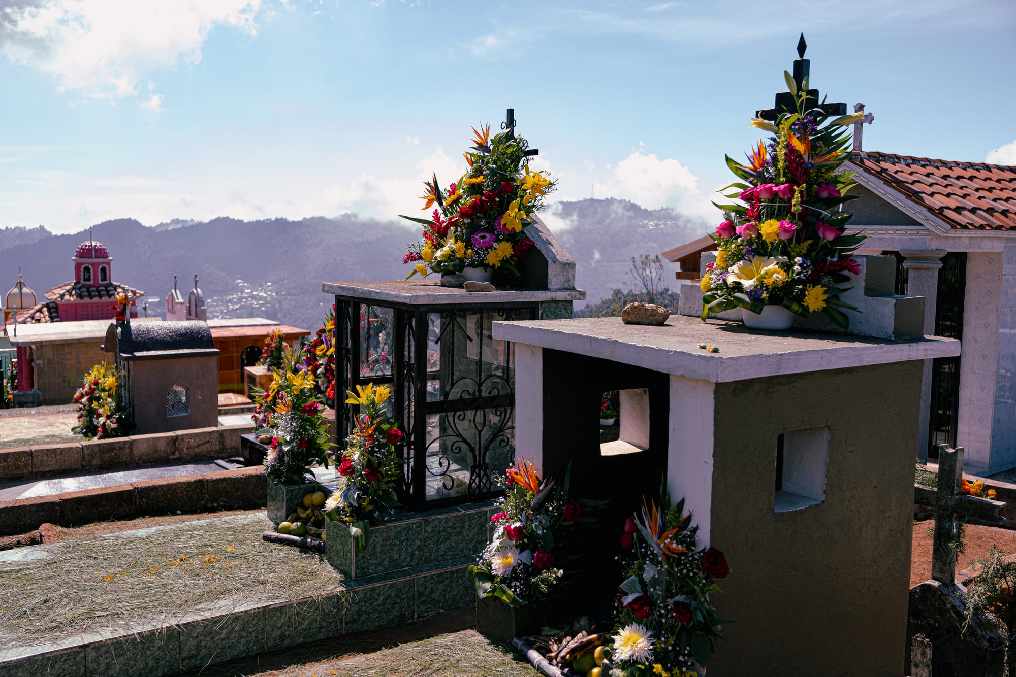 Día de Muertos at Zinacantán Cemetery in Zinacantán, Chiapas, Mexico, above ground graves and small mausoleums decorated with elaborate floral arrangements, marigolds roses lilies and tropical flowers placed on tombs, hillside cemetery with mountain views and clouds overlooking the valley near San Cristóbal de las Casas