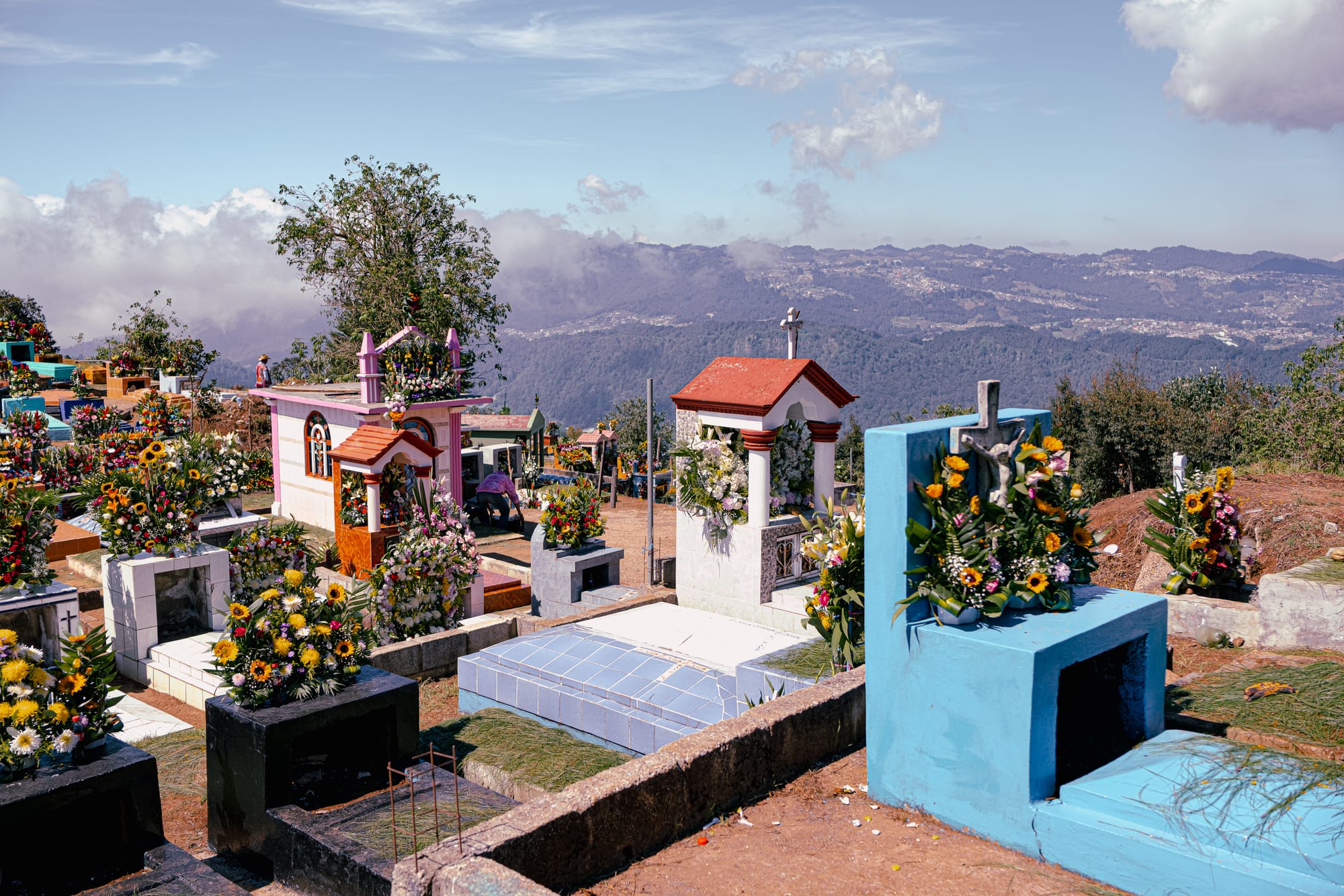 Día de Muertos at Zinacantán Cemetery in Zinacantán, Chiapas, Mexico, hillside cemetery with above ground graves and small mausoleums decorated with elaborate floral arrangements, colorful tombs overlooking mountain valleys and clouds, Day of the Dead flower traditions shaped by local flower cultivation near San Cristóbal de las Casas