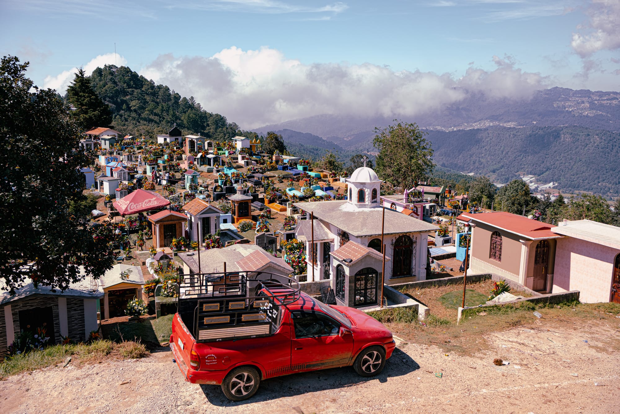 Día de Muertos at Zinacantán Cemetery in Zinacantán, Chiapas, Mexico, panoramic view of hillside cemetery with colorful above ground tombs and dense floral arrangements, mountain landscape and clouds surrounding the cemetery, pickup truck parked at the edge of the graveyard during Day of the Dead near San Cristóbal