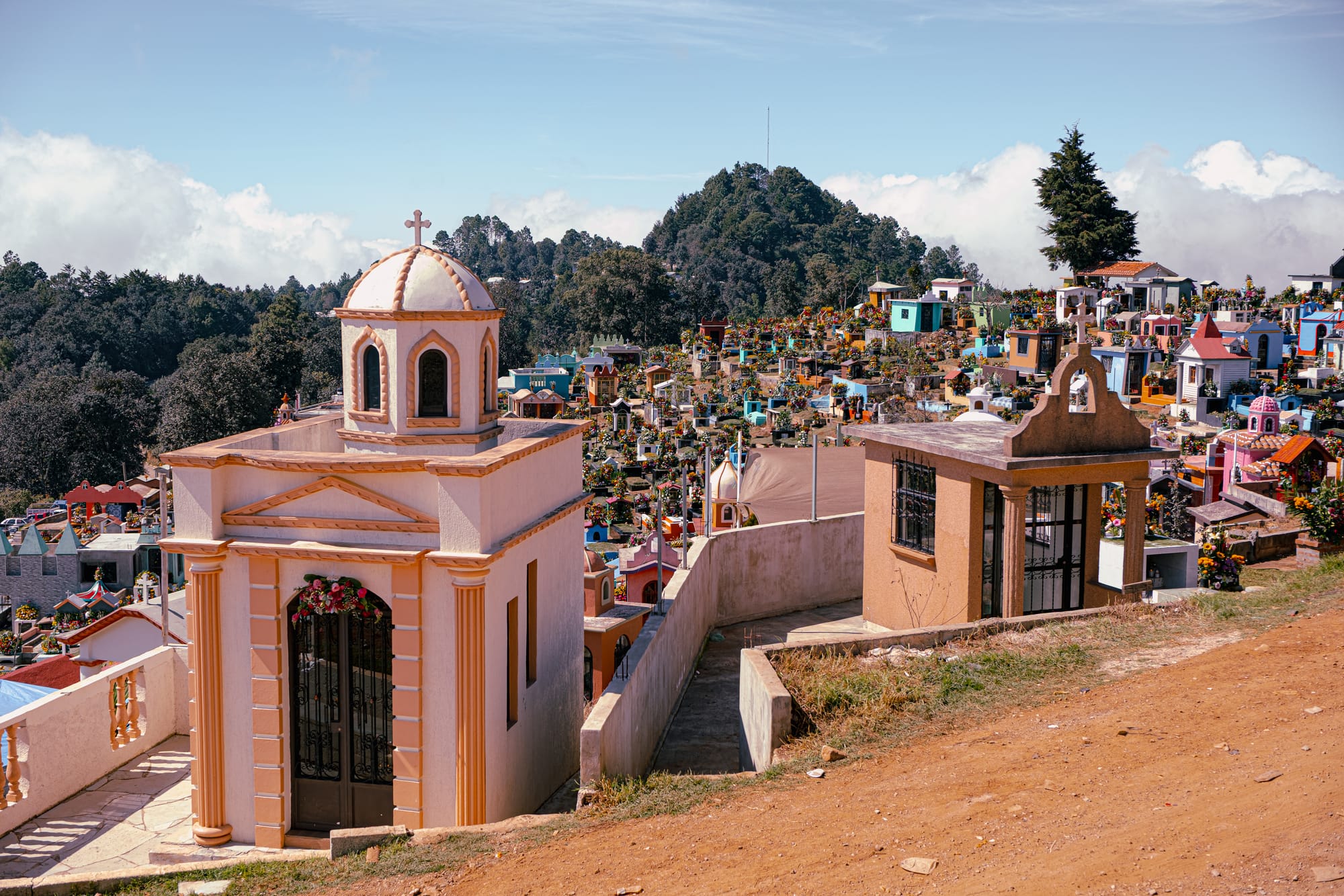 Día de Muertos at Zinacantán Cemetery in Zinacantán, Chiapas, Mexico, above ground mausoleums and chapel like tombs arranged across a hillside, colorful graves covered with floral arrangements and marigolds, mountain landscape and clouds surrounding the cemetery during Day of the Dead near San Cristóbal de las Casas