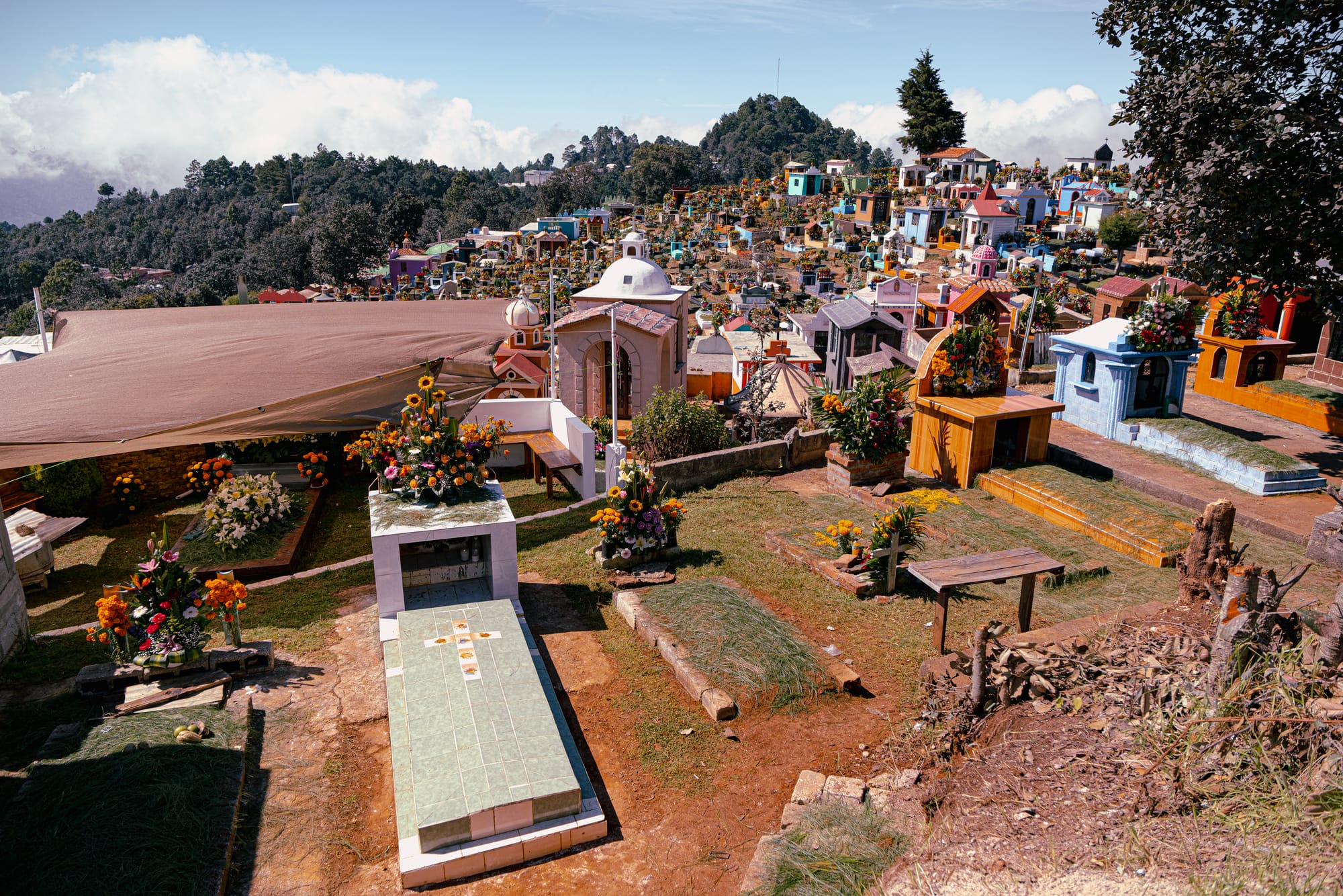 Día de Muertos at Zinacantán Cemetery in Zinacantán, Chiapas, Mexico, wide panoramic view of hillside cemetery with above ground graves and small mausoleums, floral arrangements covering tombs across multiple levels, mountain landscape and clouds surrounding the cemetery during Day of the Dead near San Cristóbal de las Casas