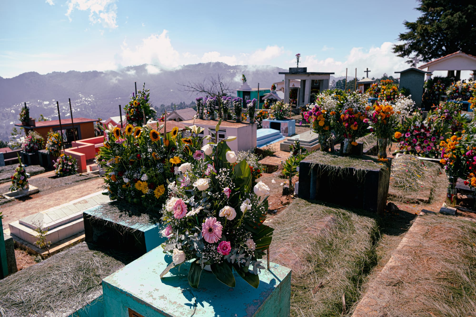 Día de Muertos at Zinacantán Cemetery in Zinacantán, Chiapas, Mexico, above ground graves arranged across a hillside and covered with dense floral arrangements, marigolds sunflowers roses and mixed flowers placed on tombs, panoramic mountain views and clouds surrounding the cemetery during Day of the Dead near San Cristóbal de las Casas