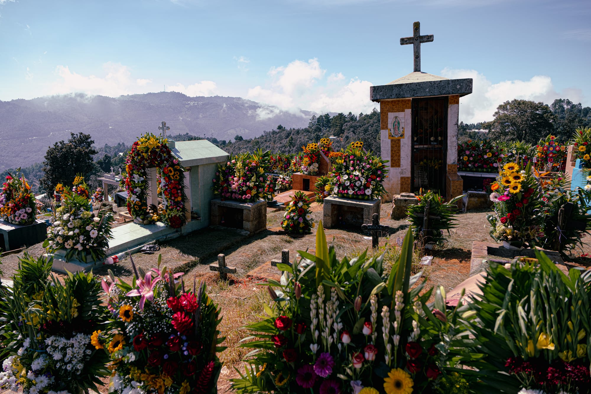 Día de Muertos at Zinacantán Cemetery in Zinacantán, Chiapas, Mexico, above ground graves and small mausoleums covered with dense floral arrangements, marigolds roses lilies and mixed flowers surrounding tombs, hillside cemetery overlooking mountains and clouds during Day of the Dead near San Cristóbal de las Casas