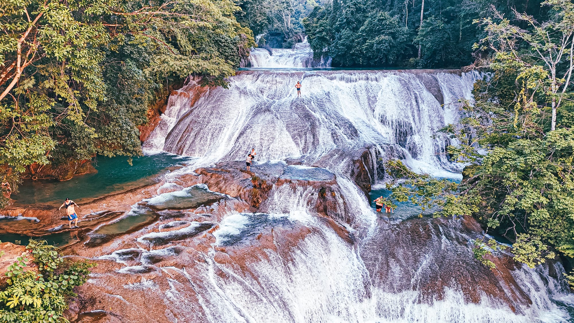 Cascadas Roberto Barrios in Chiapas, showing wide limestone waterfalls cascading in multiple tiers into turquoise pools, with people standing and swimming on the smooth rock surfaces surrounded by dense green jungle