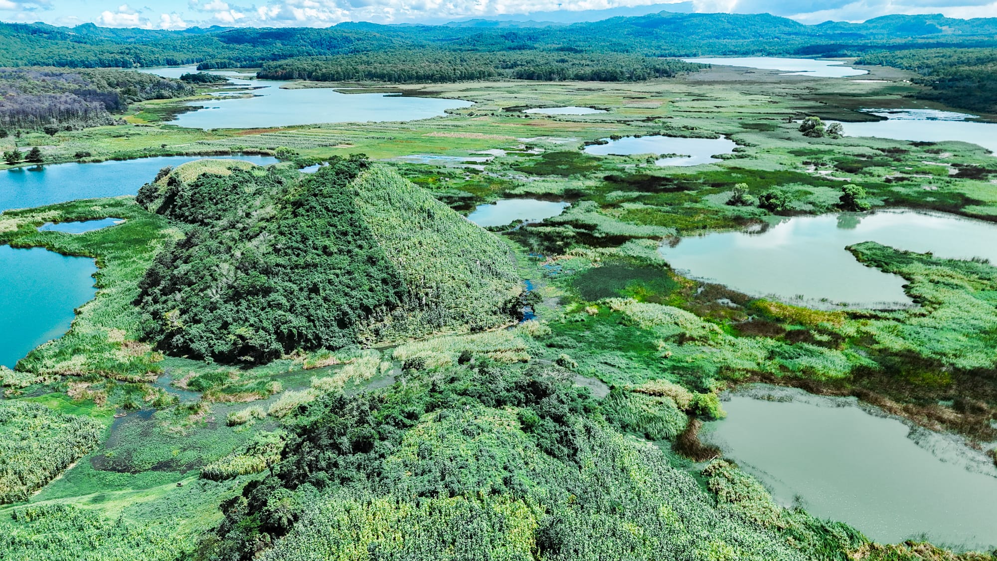 Lagunas de Montebello in Chiapas, with a patchwork of shallow lakes in varying shades of blue and green spread across wetlands and forested hills, viewed from above with rolling mountains in the distance under a clear sky