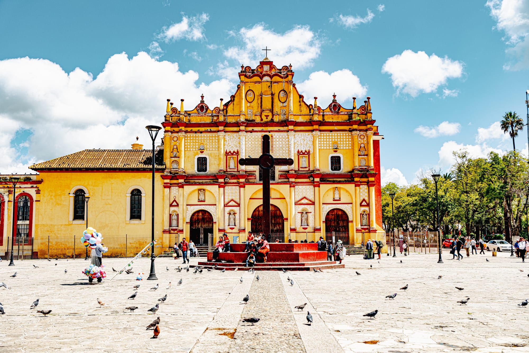 San Cristóbal Cathedral (Catedral de San Cristóbal Mártir) in the main plaza of San Cristóbal de las Casas, with its yellow baroque façade facing an open stone square where people sit, walk, and gather as pigeons scatter across the plaza under a bright blue sky